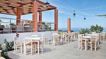 An outdoor patio with white tables and chairs, a pergola structure, and a view of the sea.
