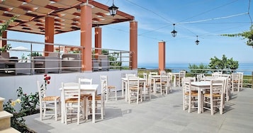 Un patio exterior con mesas y sillas blancas, una estructura de pérgola y vista al mar.