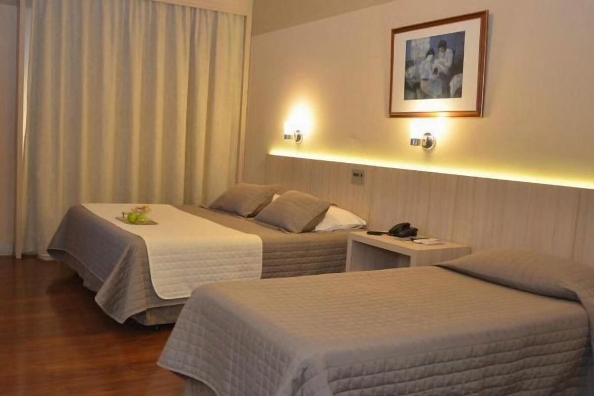 Habitación de hotel con dos camas, una mesita de noche con lámpara, un cuadro enmarcado en la pared y una ventana con cortinas.