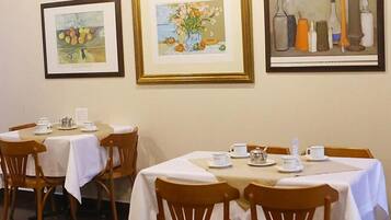 A dining area with tables set for two, framed artwork on the wall, and wooden chairs.