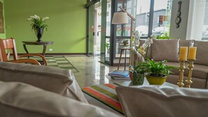 A modern living room with a green wall, a sofa, a coffee table with books and plants, a lamp, and a view of the street through large windows. - Village Hotel (Ponta Grossa)