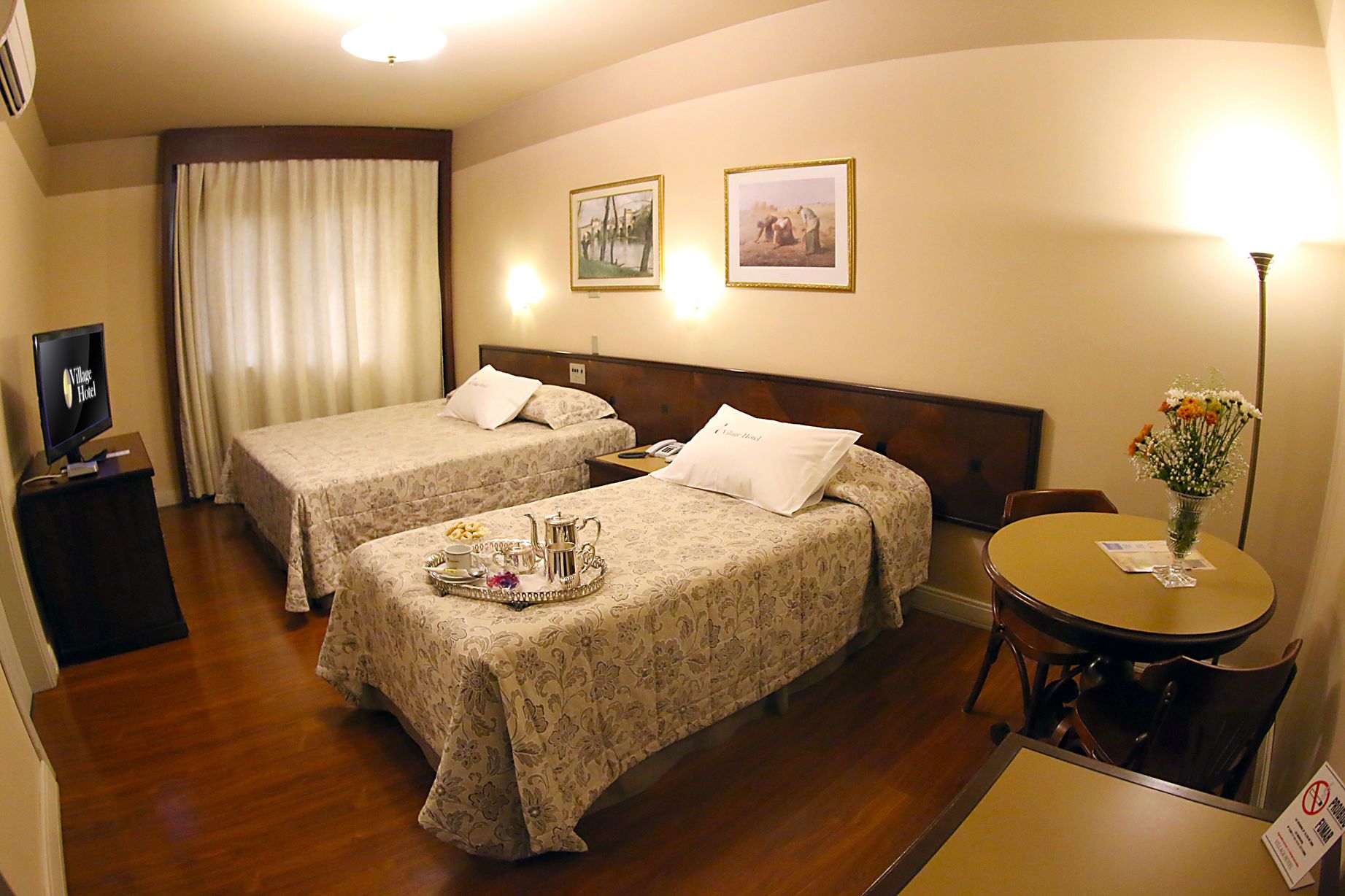 Ein Hotelzimmer mit zwei Betten, einem kleinen Tisch mit Blumenvase und einem Fernseher auf einem Schrank.