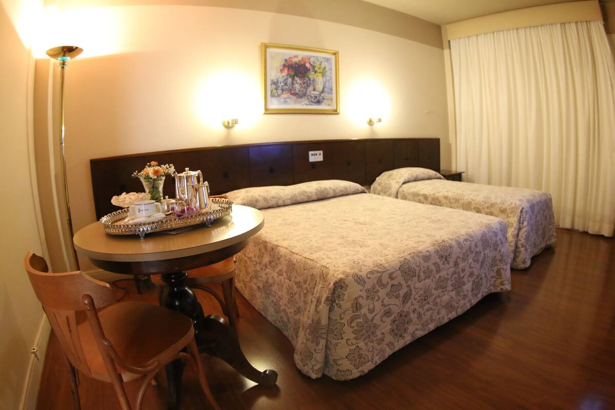 A hotel room with two beds, a dining table with a tray of breakfast items, a chair, and a painting on the wall.