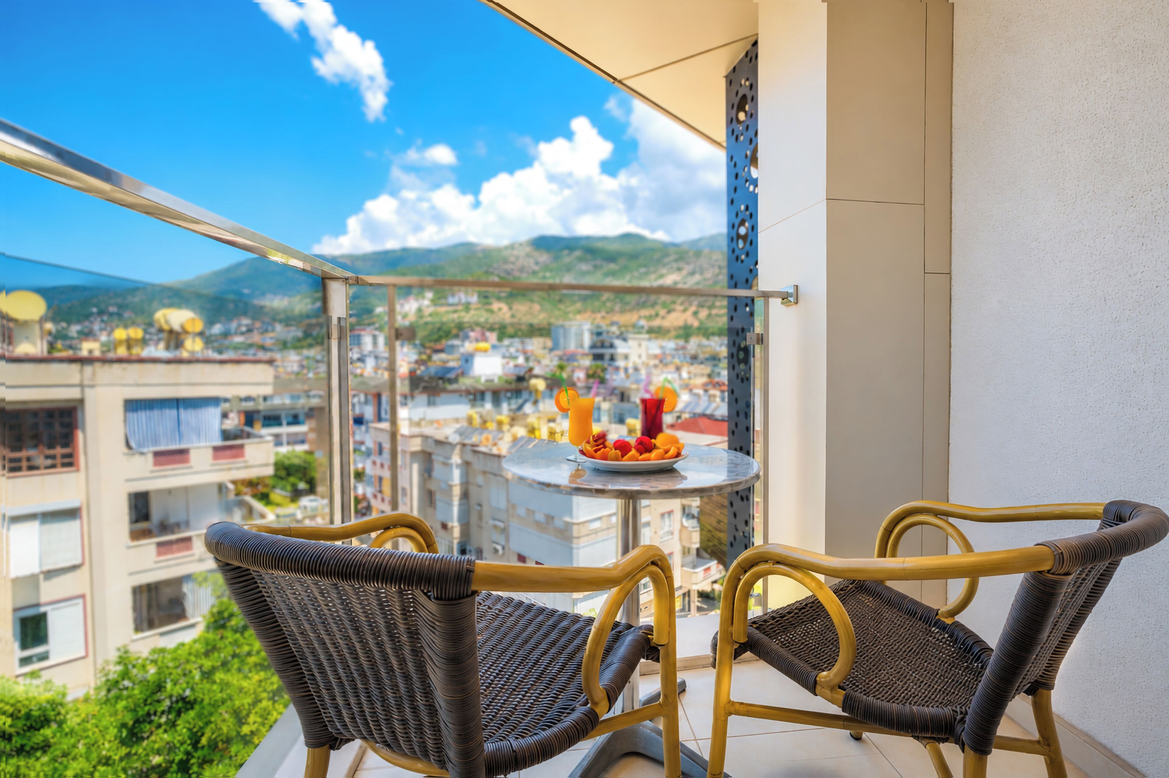 Een balkon met rieten stoelen, een kleine tafel met fruit en een drankje, en uitzicht op de stad.
