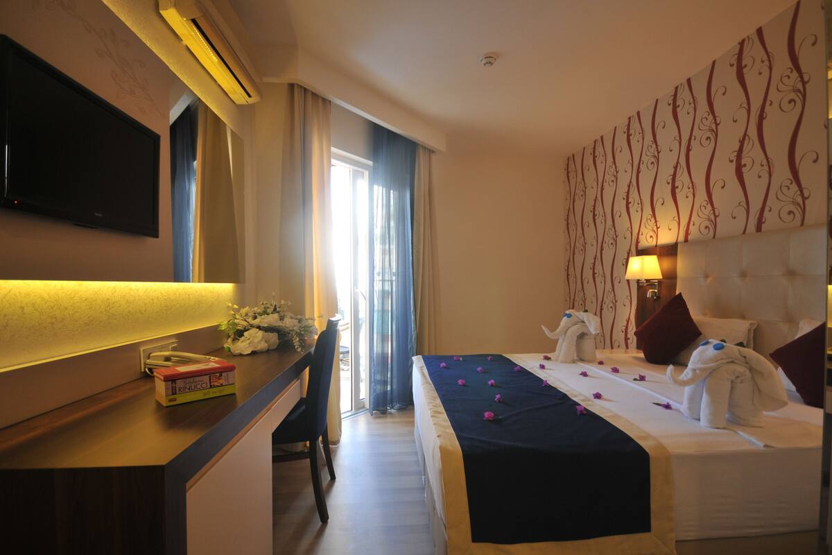 Una habitación de hotel con cama, escritorio con teléfono, televisor y ventana con cortinas.