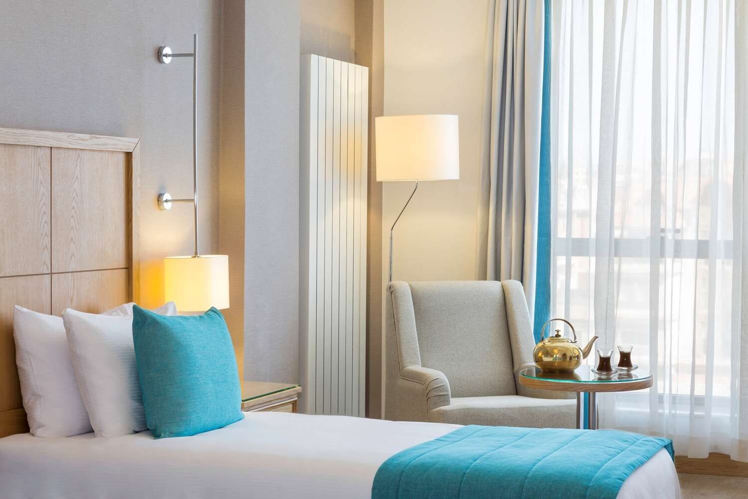 A hotel room with a bed, a chair, a table with a teapot, and a lamp.