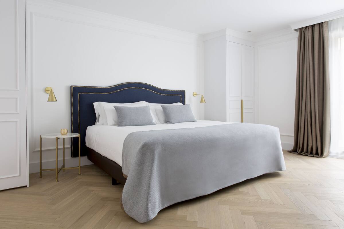 Un lit bien fait, avec une tête de lit sombre, une table de chevet avec une lampe et une vue sur une fenêtre avec des rideaux.