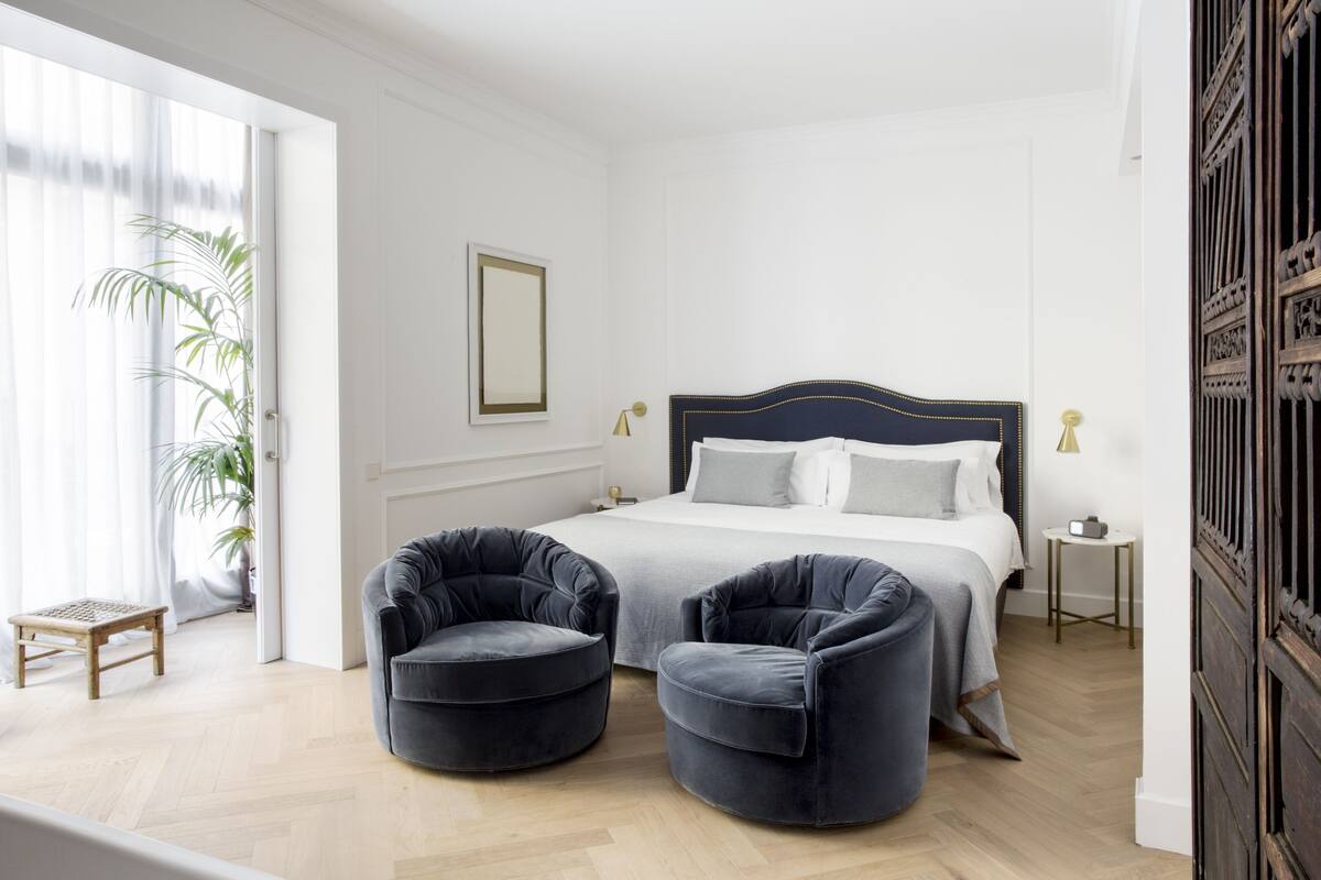 Une chambre à coucher avec un grand lit, deux fauteuils, une table de chevet, une lampe et une plante en pot.