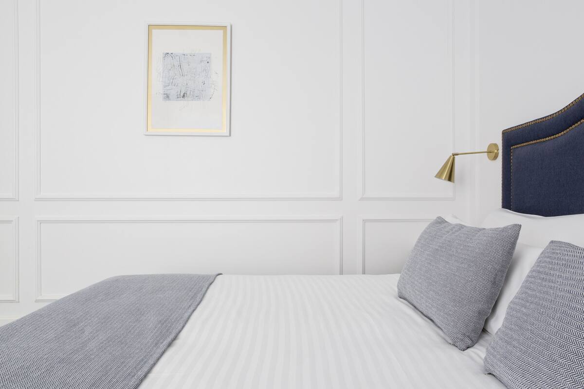 Un lit bien fait, recouvert d’une couvre-lit à rayures, d’un plaid gris et de deux coussins à motifs. Une œuvre d’art encadrée est accrochée au mur au-dessus du lit.