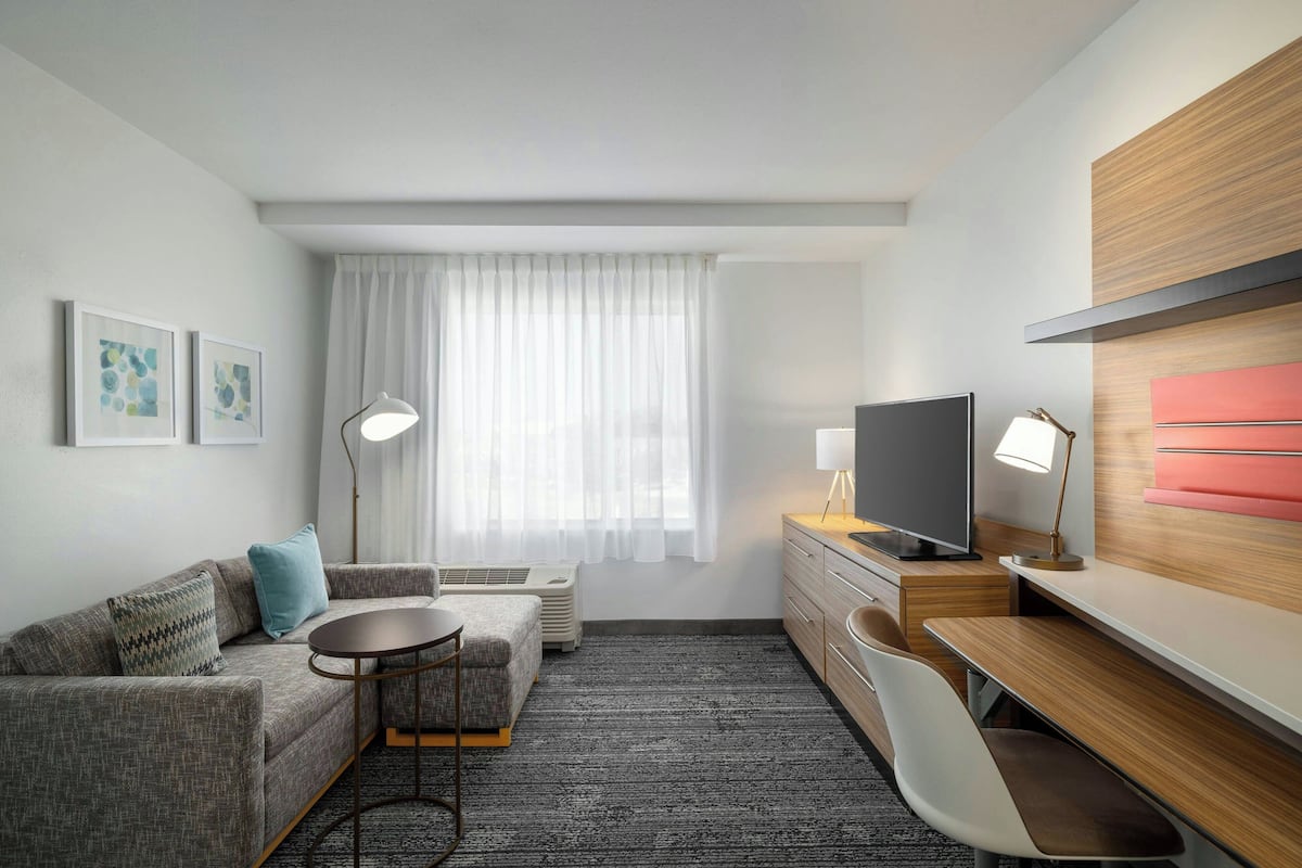 Een moderne hotelkamer met een bank, een klein rond tafeltje, een bureau met een stoel, een televisie en een groot raam met dunne gordijnen.