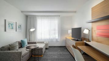 Una habitación de hotel moderna con sofá, una pequeña mesa redonda, un escritorio con silla, televisor y un amplio ventanal con cortinas translúcidas.