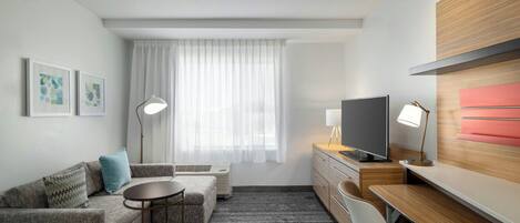 Una habitación de hotel moderna con sofá, una pequeña mesa redonda, un escritorio con silla, televisor y un amplio ventanal con cortinas translúcidas.