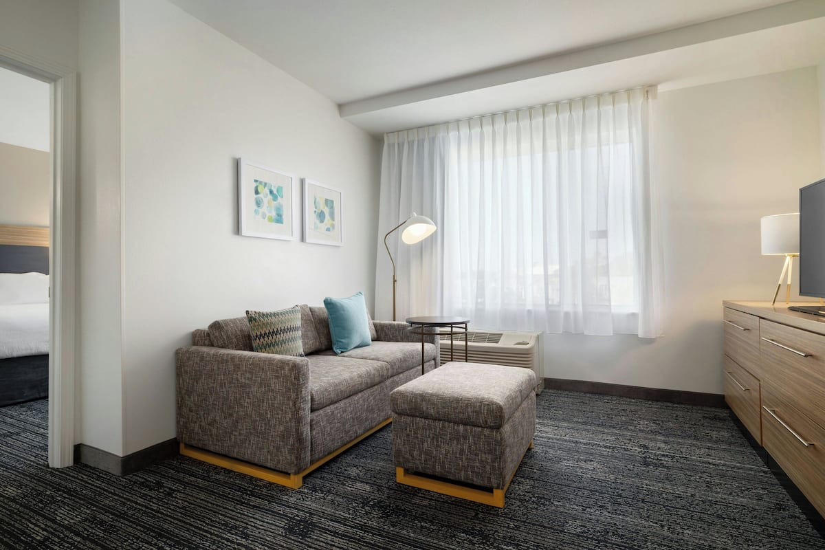 Een hotelkamer met een grijze bank, een bijpassend voetenbankje, een tafeltje, een lamp, een televisie en een raam met dunne gordijnen.
