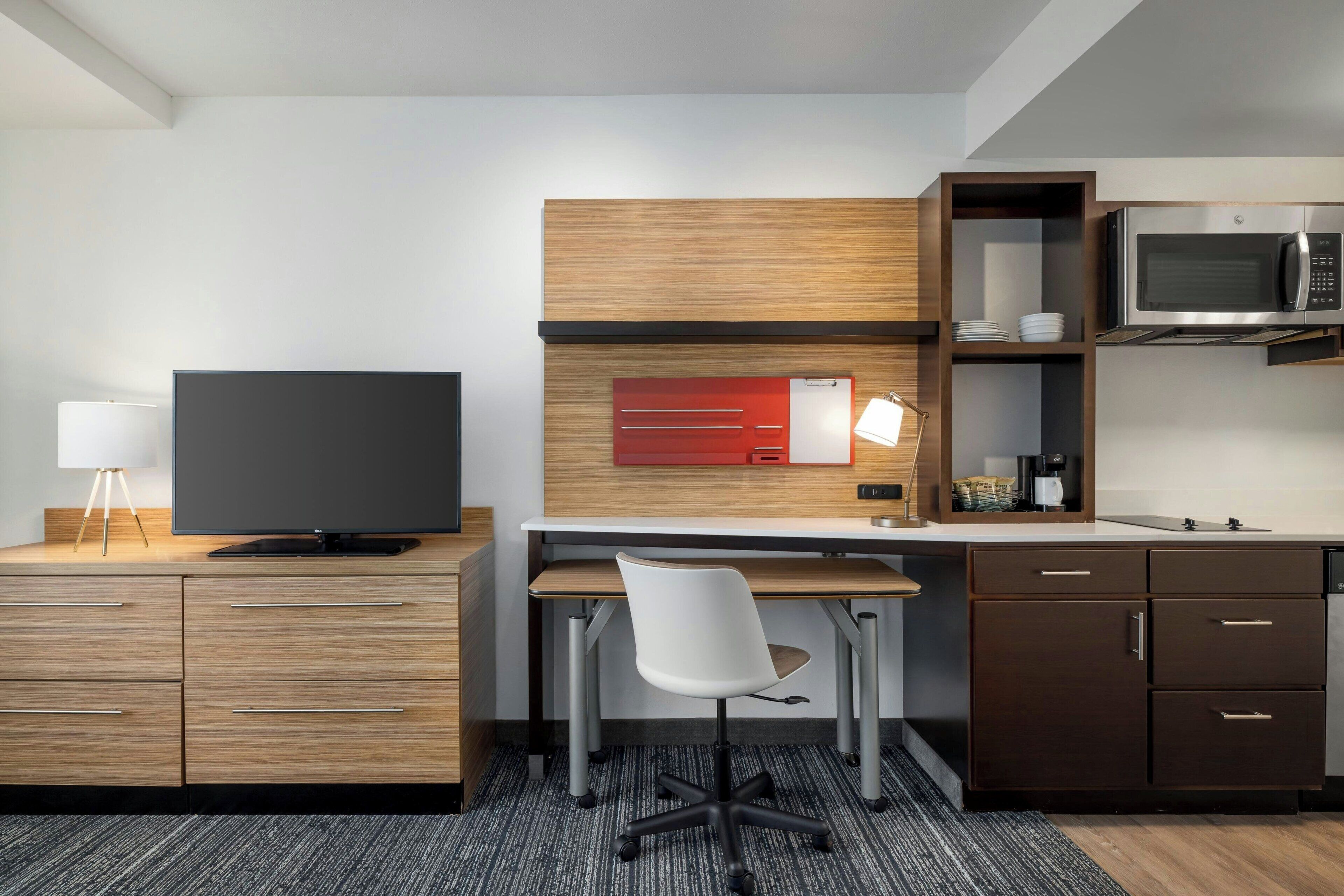 Una habitación de hotel moderna con un escritorio de madera, un televisor de pantalla plana, una silla de oficina blanca, un microondas y una pequeña cocina.