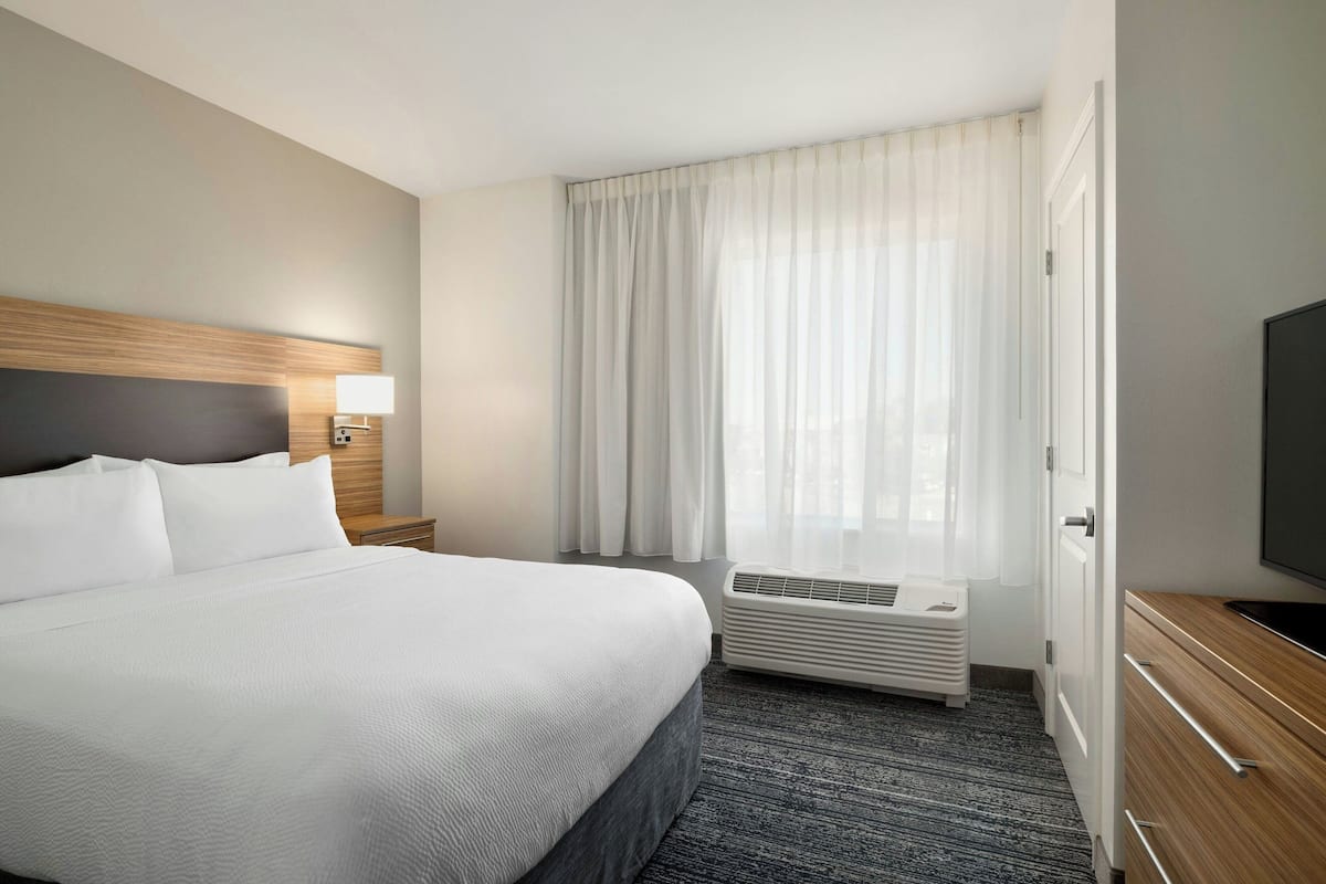 Habitación de hotel con una cama grande, una mesita de noche con lámpara, una ventana con cortinas, un televisor montado en la pared y un calentador incorporado.