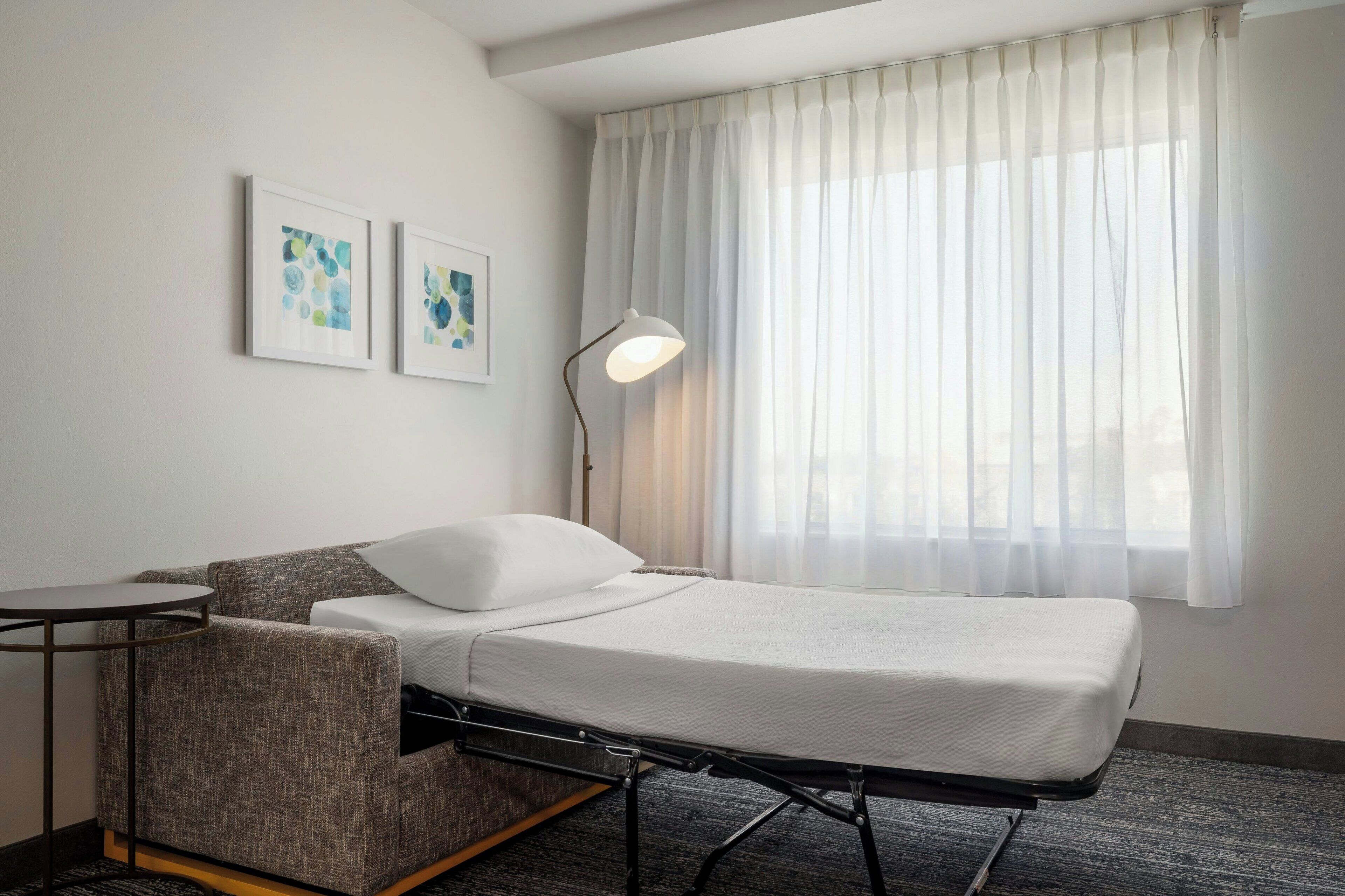 Ein Hotelzimmer mit einem Bett, einem kleinen Tisch, einem Stuhl, einer Lampe und zwei gerahmten Kunstwerken an der Wand.