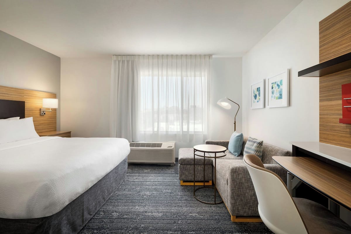 Una habitación de hotel moderna con una cama grande, un sofá, una pequeña mesa redonda y un escritorio. Se aprecia una ventana con cortinas translúcidas, una lámpara de pared y dos cuadros enmarcados.
