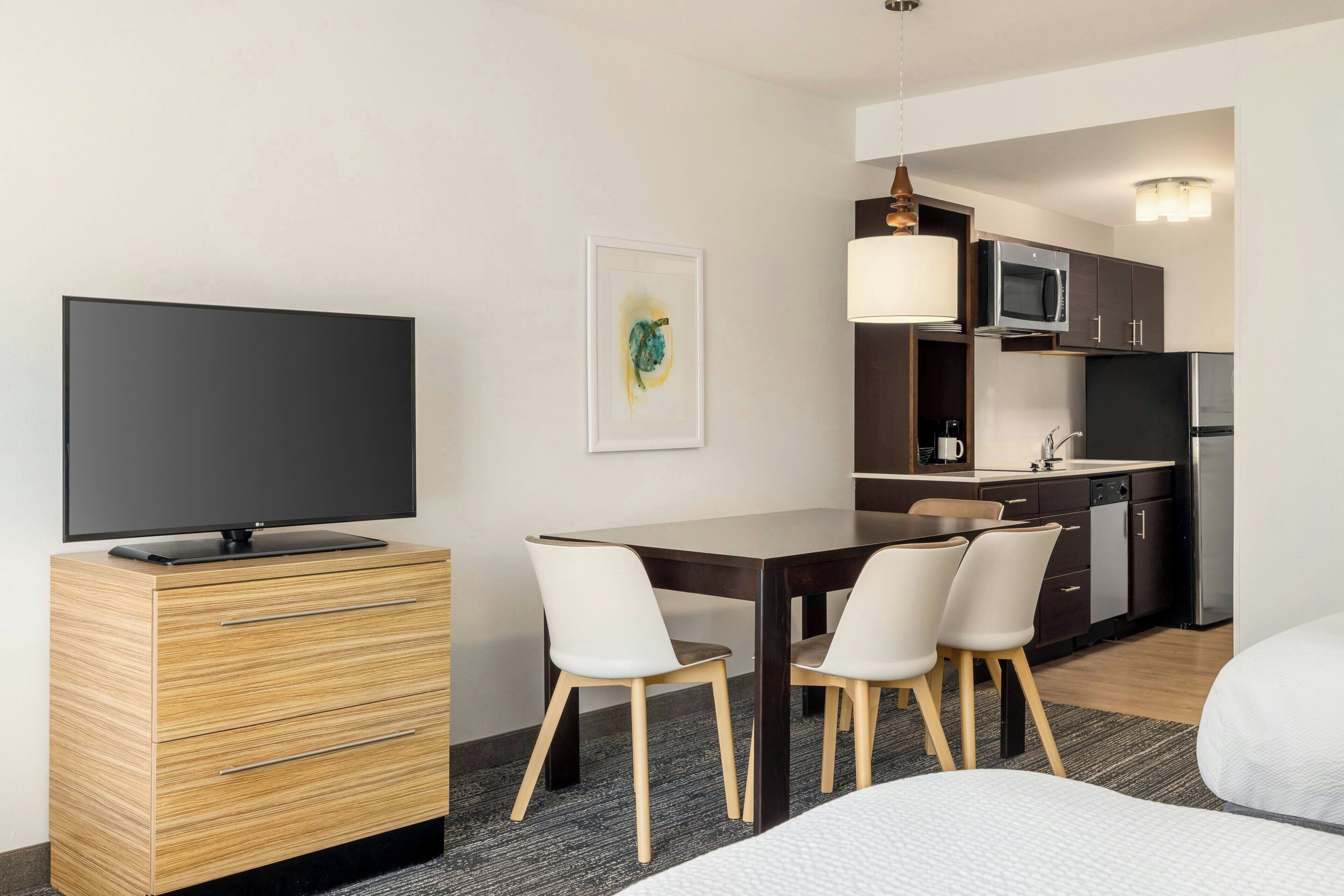 A modern hotel room with a flat-screen TV, a wooden dresser, a dining table with chairs, a kitchen area with a microwave, a refrigerator, and a hanging light fixture.