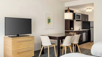 A modern hotel room with a flat-screen TV, a wooden dresser, a dining table with chairs, a kitchen area with a microwave, a refrigerator, and a hanging light fixture.