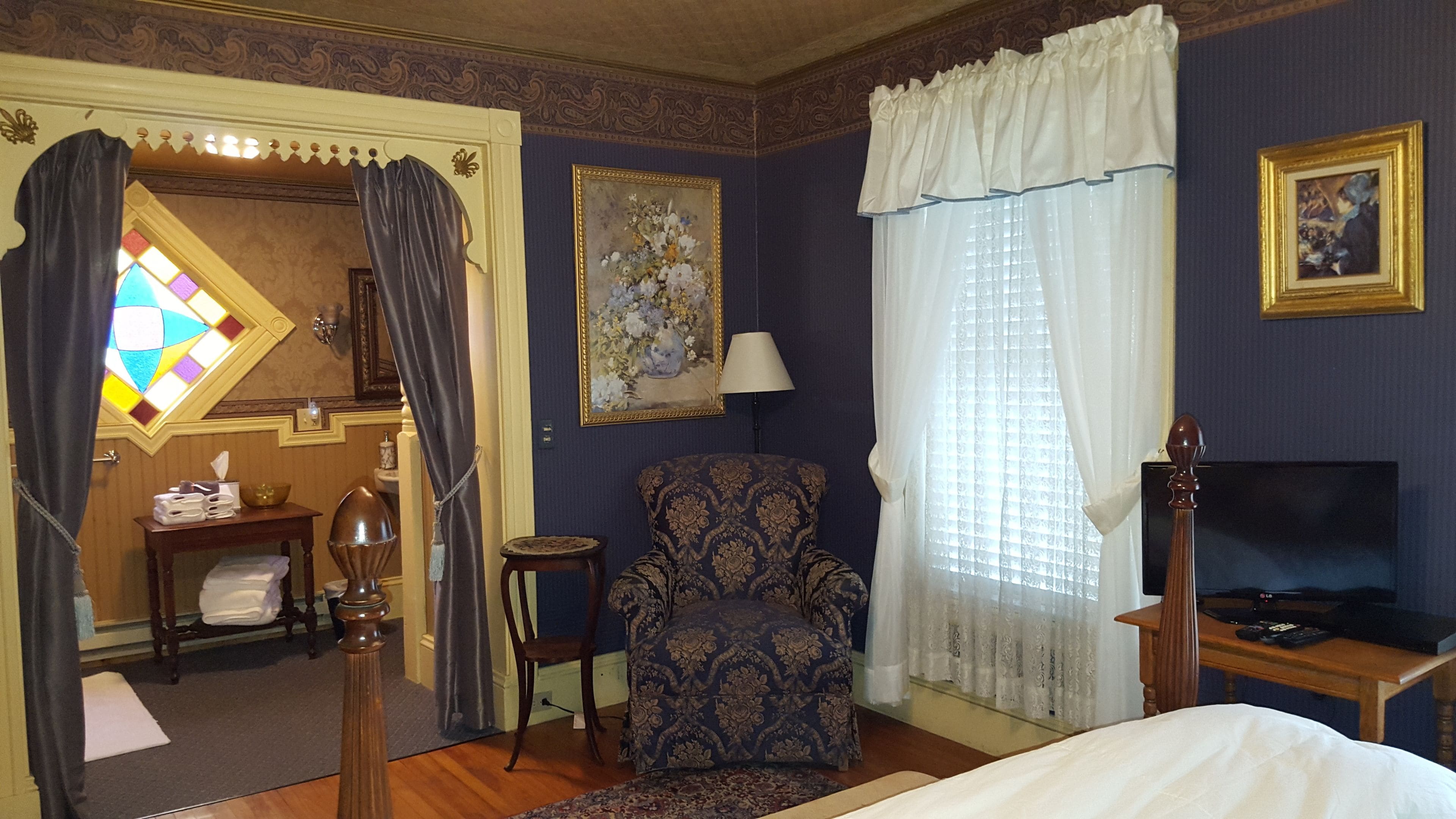 Habitación de hotel con cama, una silla, televisor, lámpara, un cuadro y un vitral.