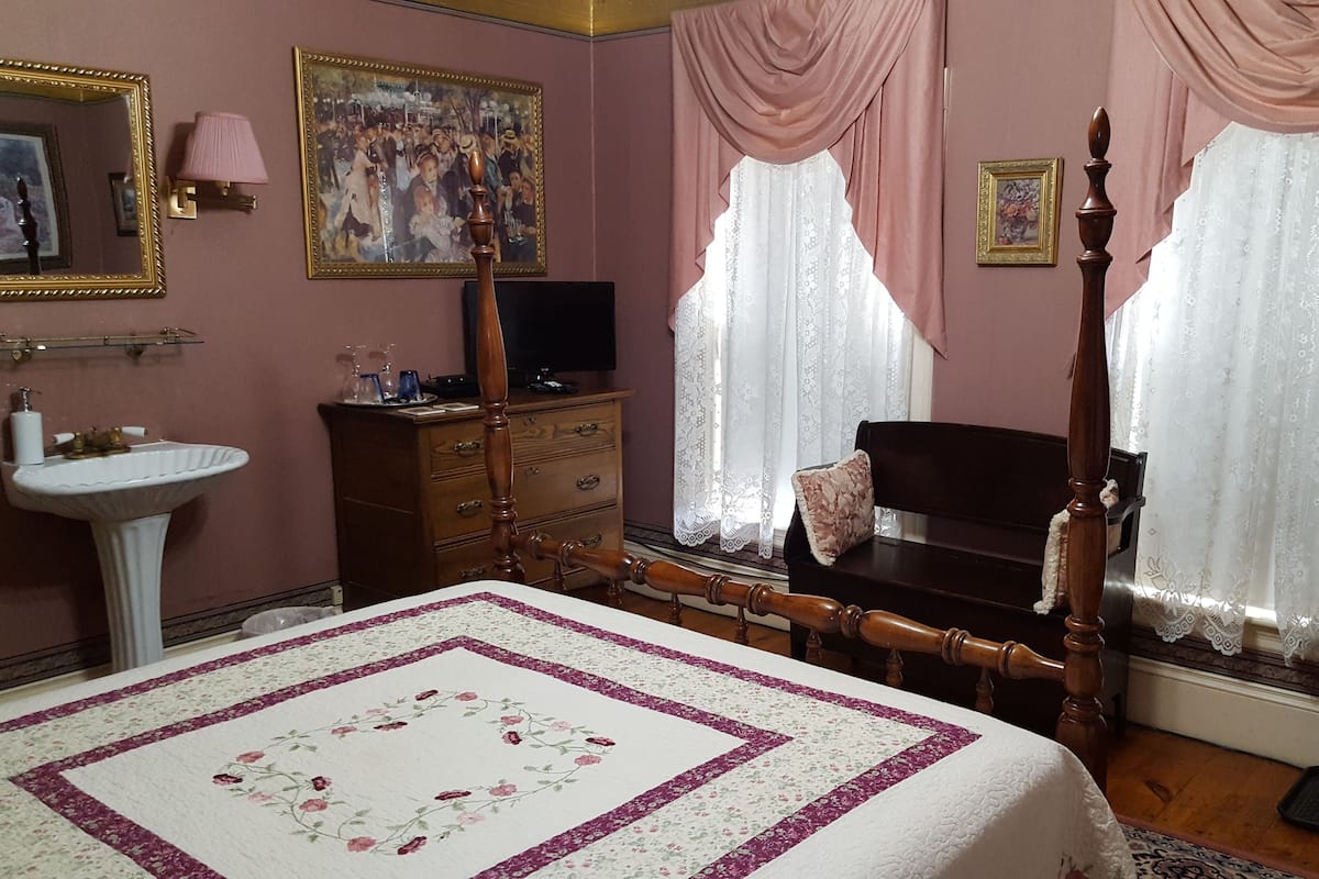 Un dormitorio con cama, tocador, espejo, televisor y dos ventanas con cortinas translúcidas.