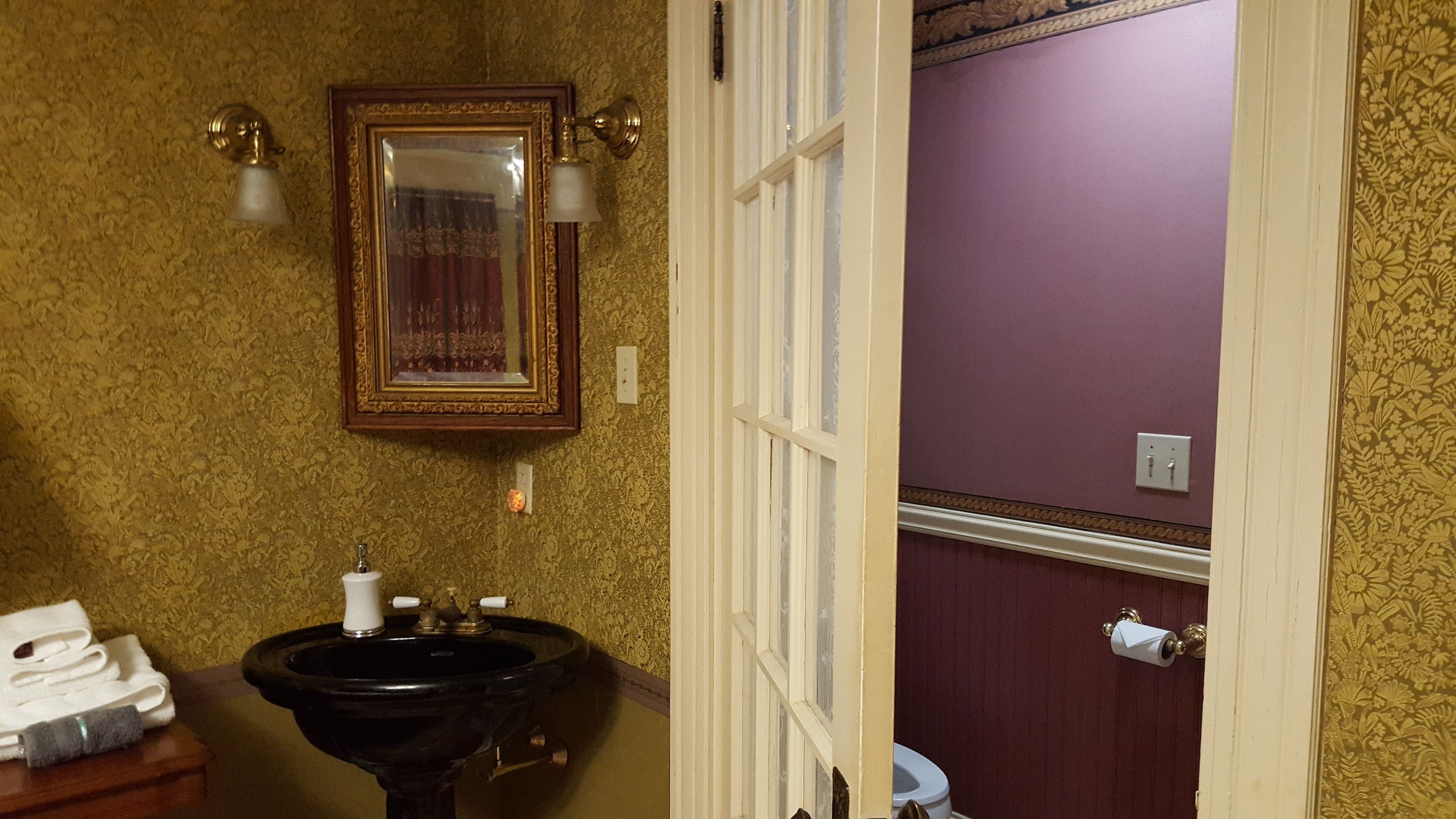 Un baño con un lavamanos negro, un espejo con marco dorado y un inodoro con pared morada.