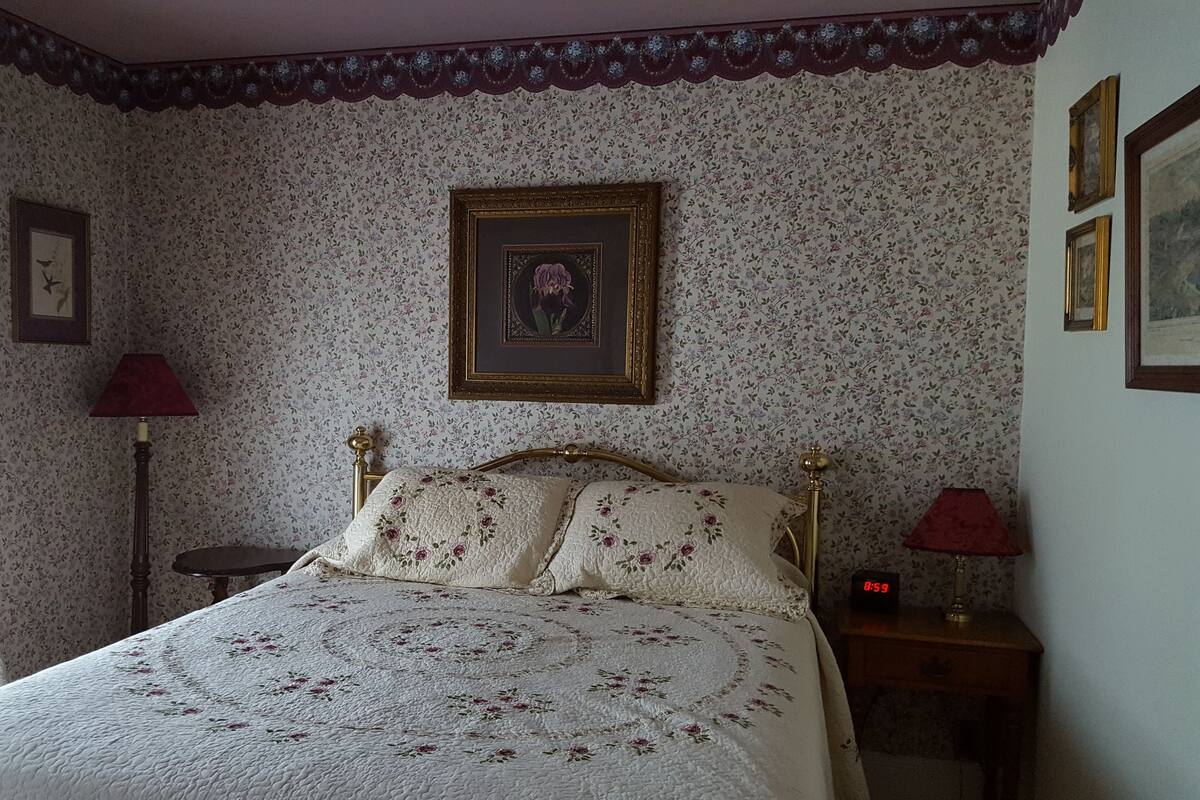 Un dormitorio con papel tapiz floral, una cama con colcha, una mesita de noche con lámpara y un reloj de pared.