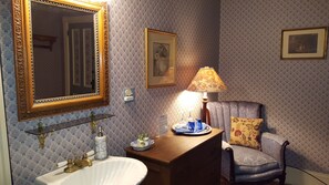A bathroom with a large ornate mirror, a sink with a gold faucet, a vanity with a mirror, a floral lamp, and a chair with a cushion.
