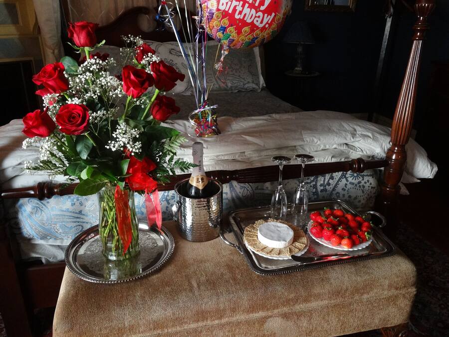 Une chambre à coucher avec un lit, une table de chevet ornée d’un vase de roses rouges, une bouteille de champagne, deux verres à vin, un plateau avec des fraises et un dessert.