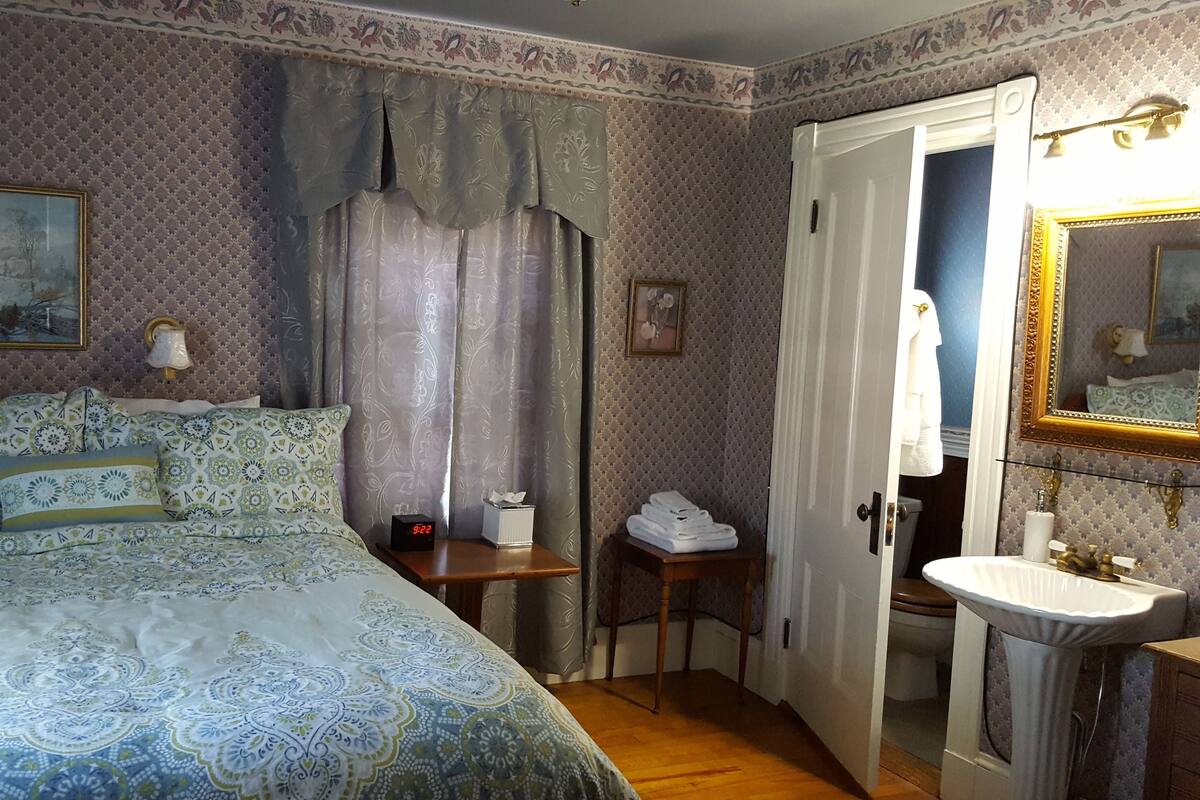 Un dormitorio con cama, mesita de noche, tocador, espejo, puerta y baño con lavamanos.