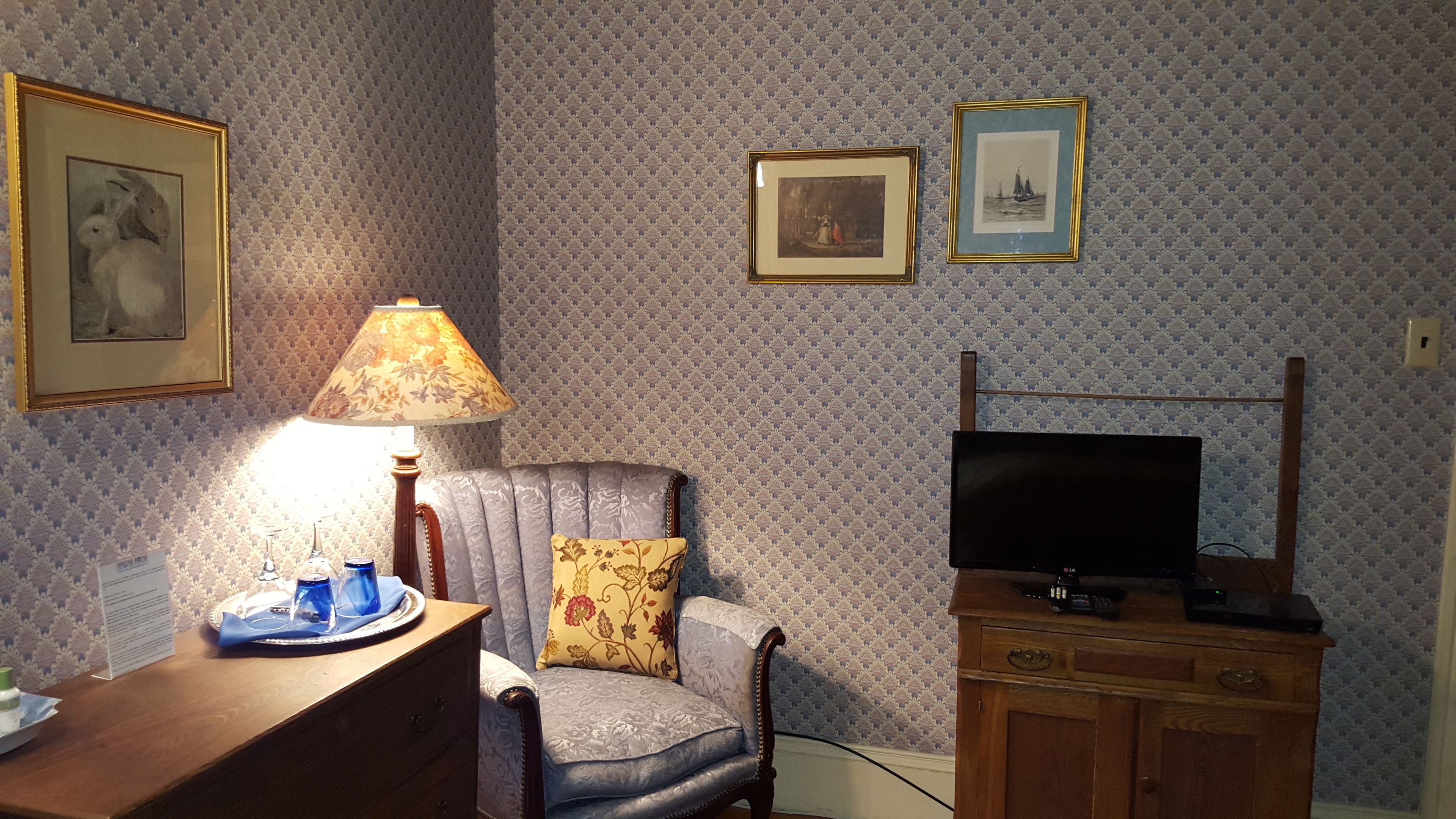 A room with patterned wallpaper, a wooden desk with a lamp, a cushioned armchair, a television on a wooden stand, and framed pictures on the wall.