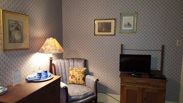 A room with patterned wallpaper, a wooden desk with a lamp, a cushioned armchair, a television on a wooden stand, and framed pictures on the wall.