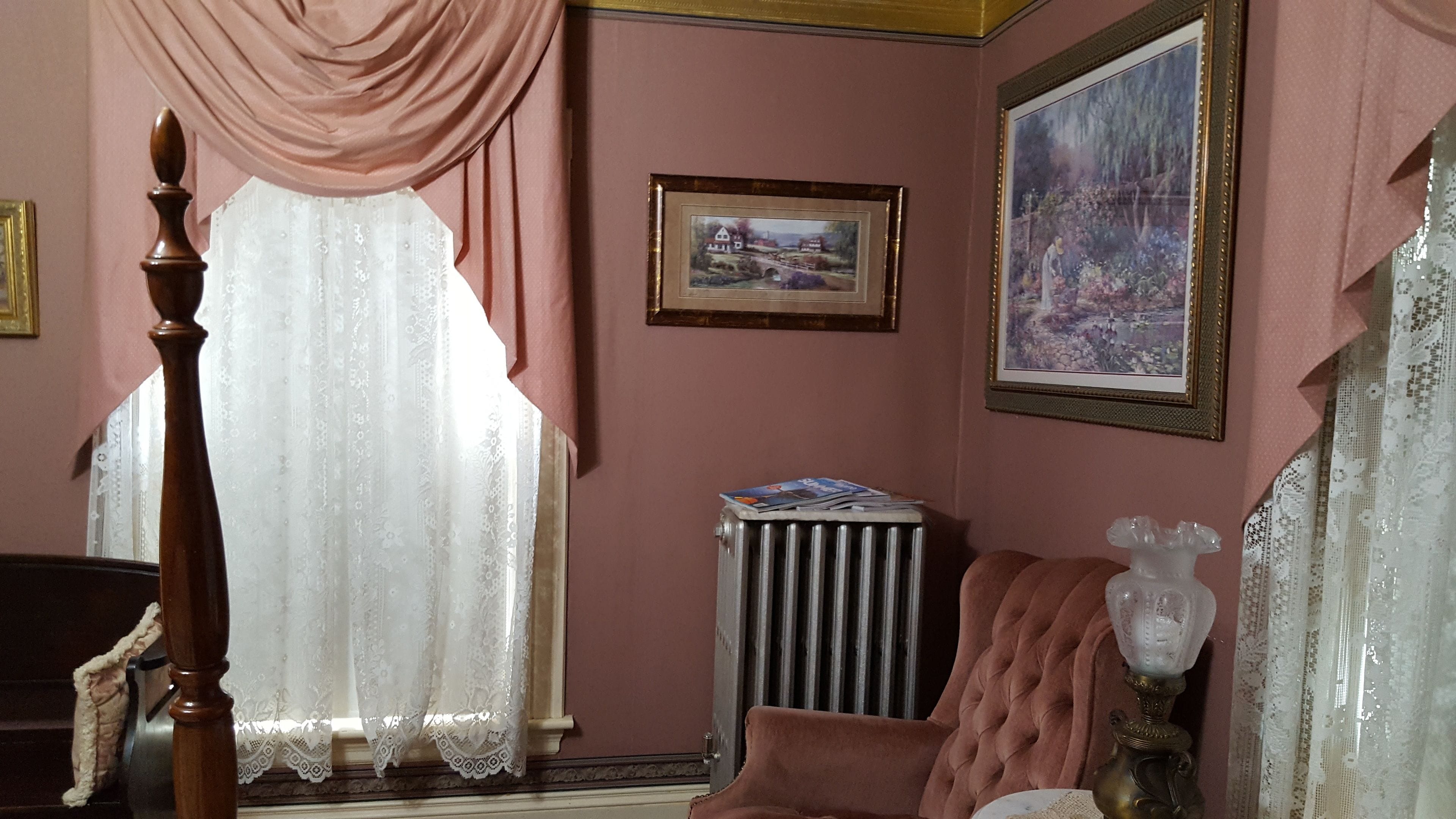 Una habitación con una cama con dosel de madera, un radiador, un cuadro de paisaje enmarco y un florero sobre una mesa.