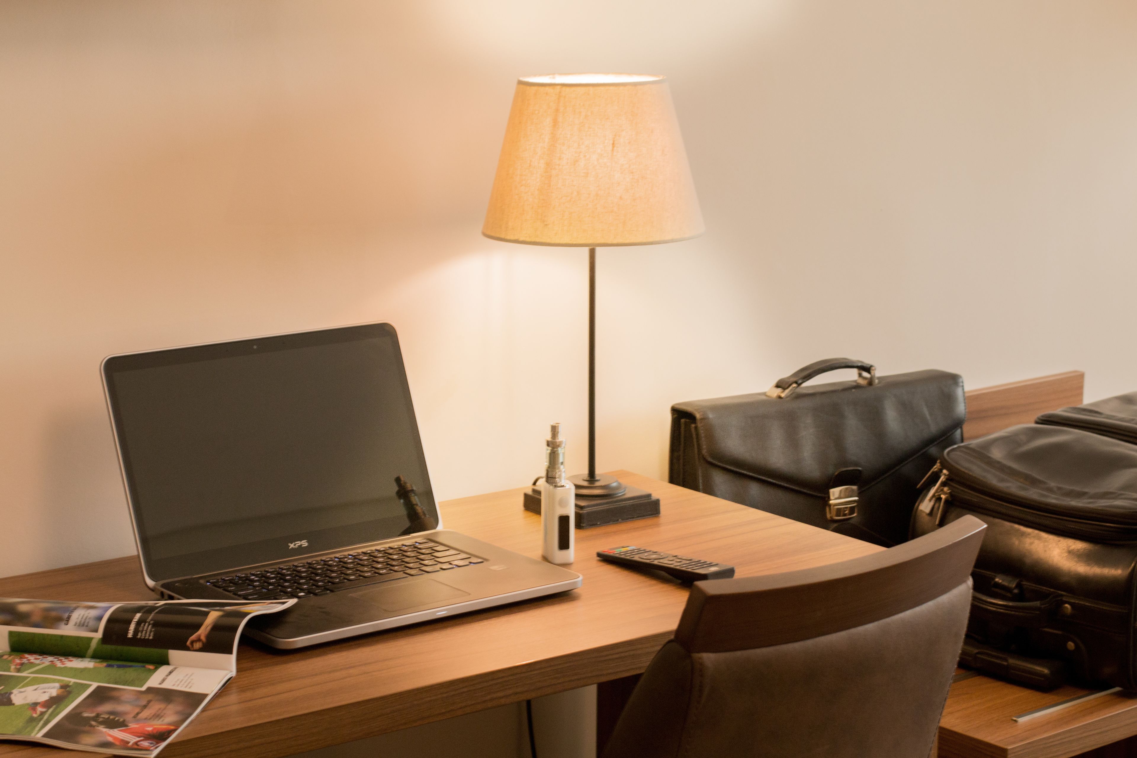 Une chambre d’hôtel avec un bureau en bois, un ordinateur portable, une lampe, une télécommande et un sac en cuir.