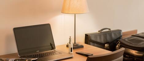 Ein Hotelzimmer mit einem Holztisch, einem Laptop, einer Lampe, einer Fernbedienung und einer Lederhandtasche.