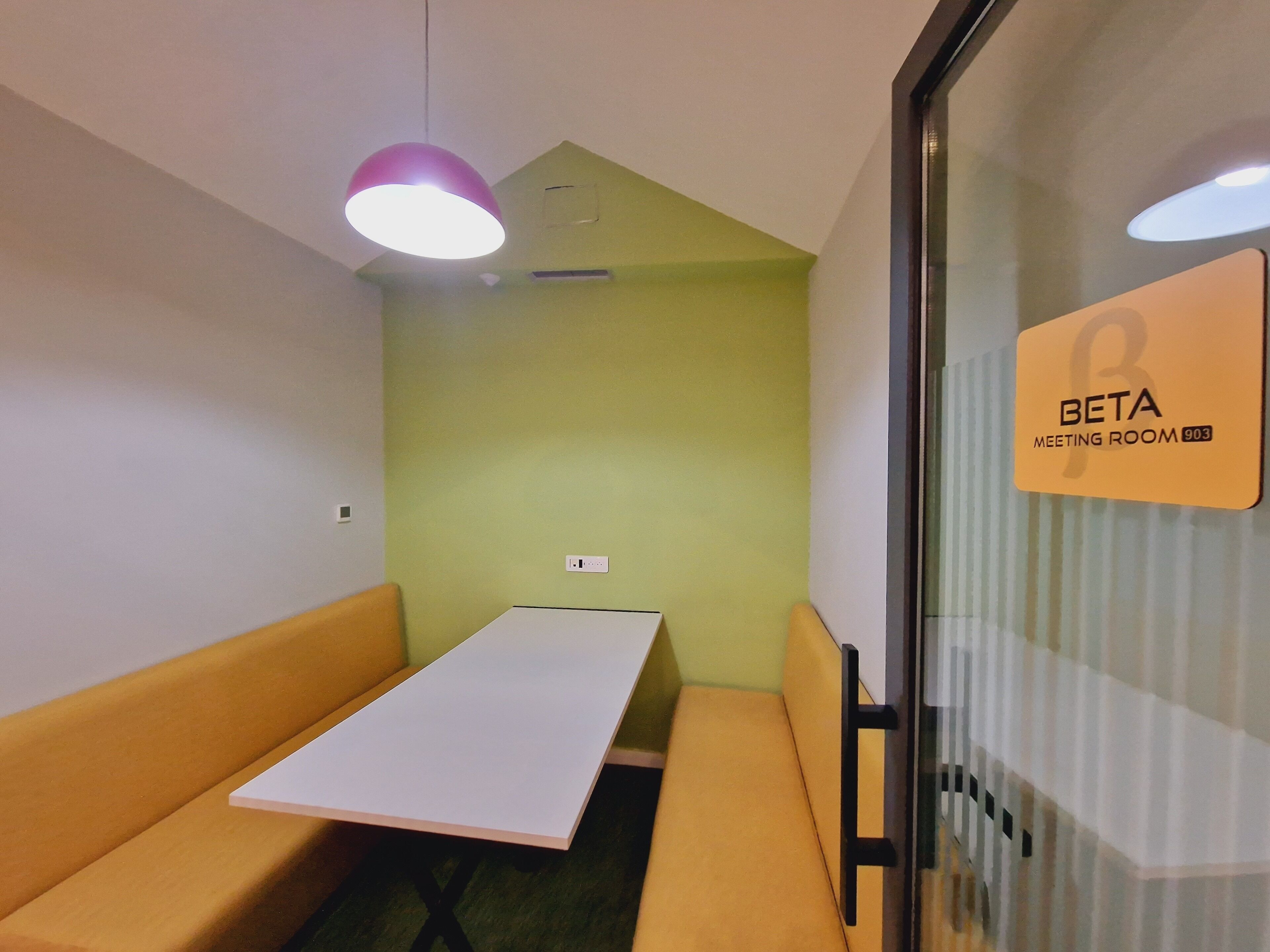 Una sala de reuniones con una mesa blanca, asientos acolchados amarillos, un separador de vidrio y un letrero que indica que es la sala 903.