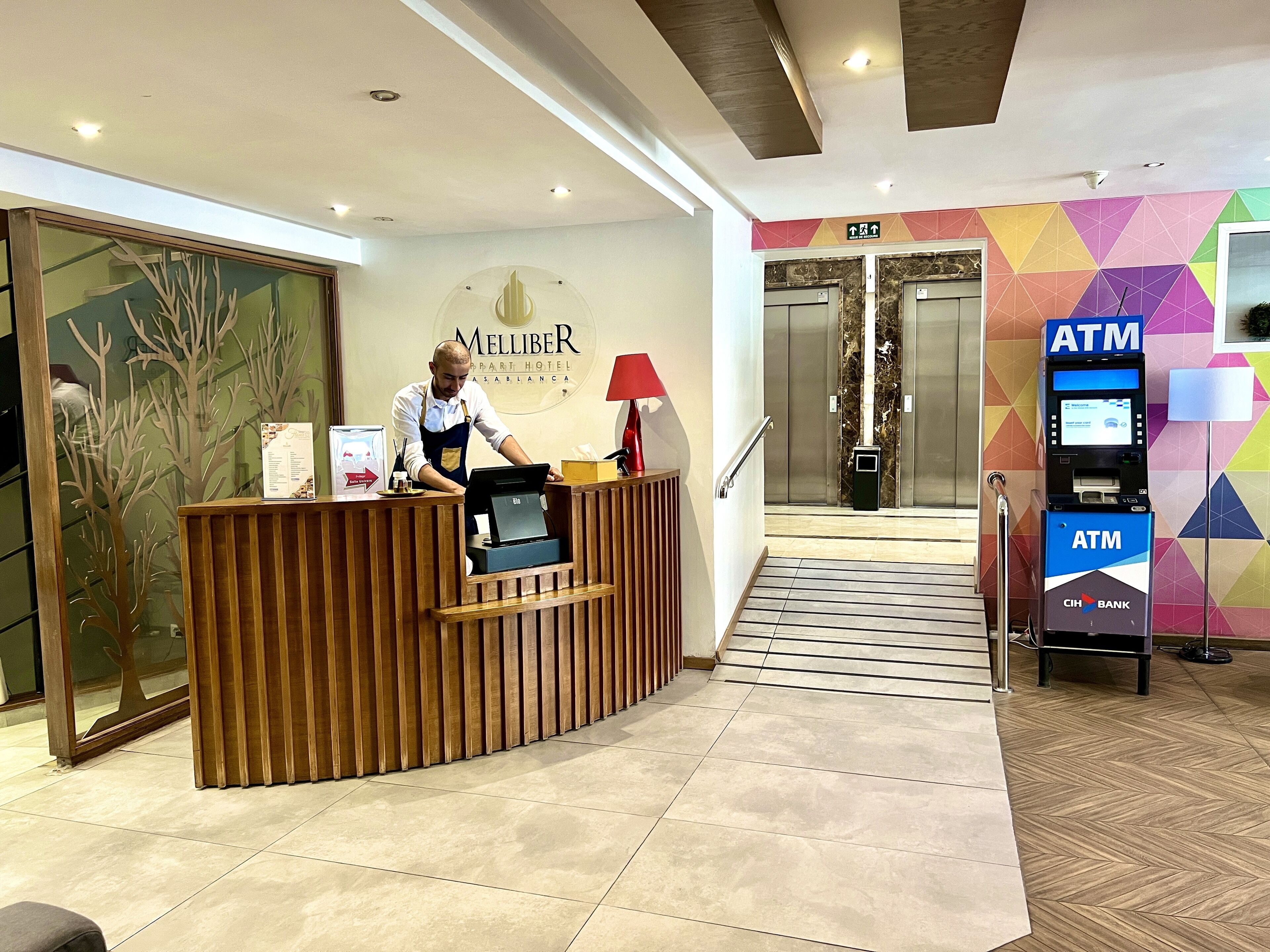 A hotel reception desk with a receptionist, an ATM, and a staircase leading to elevators.