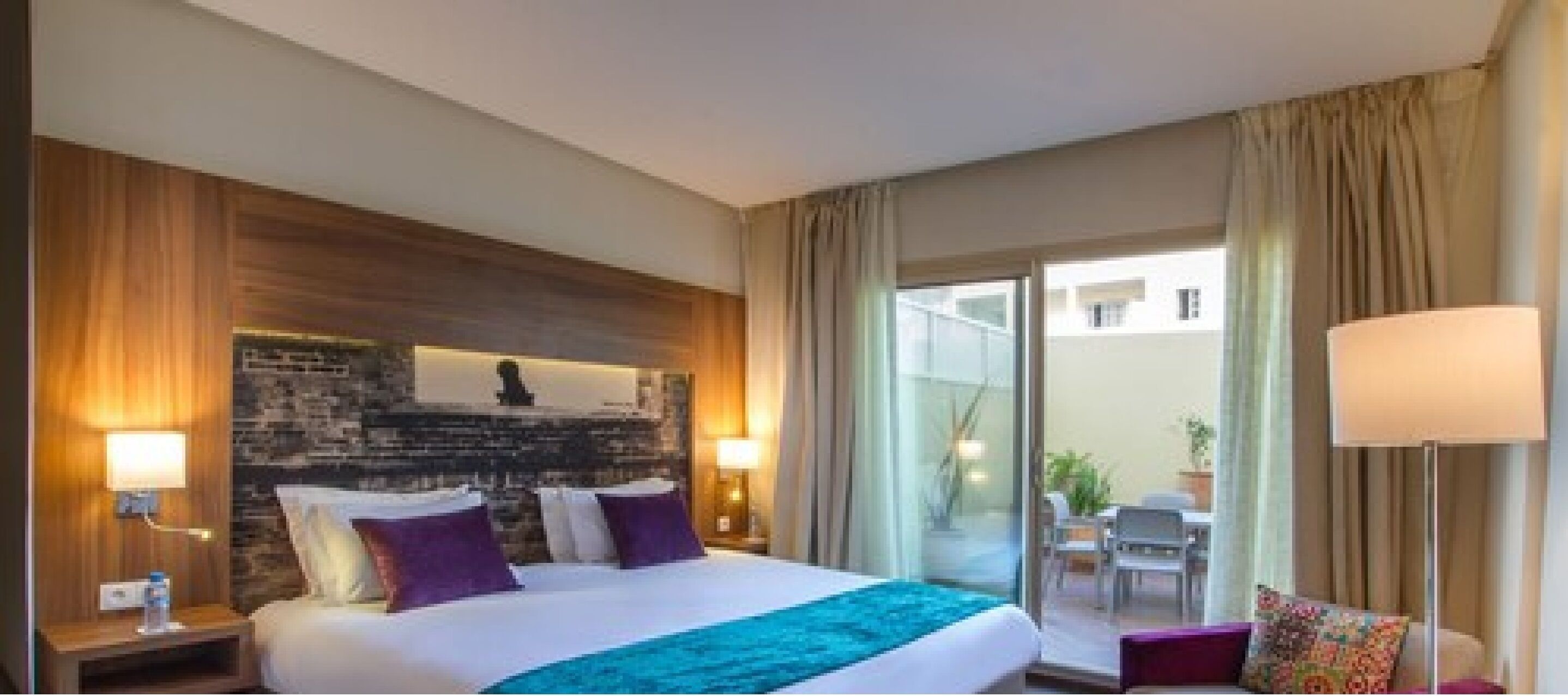 Habitación de hotel con una cama grande, una pared de acento de piedra, vistas al exterior y una zona de estar con mesa y sillas.