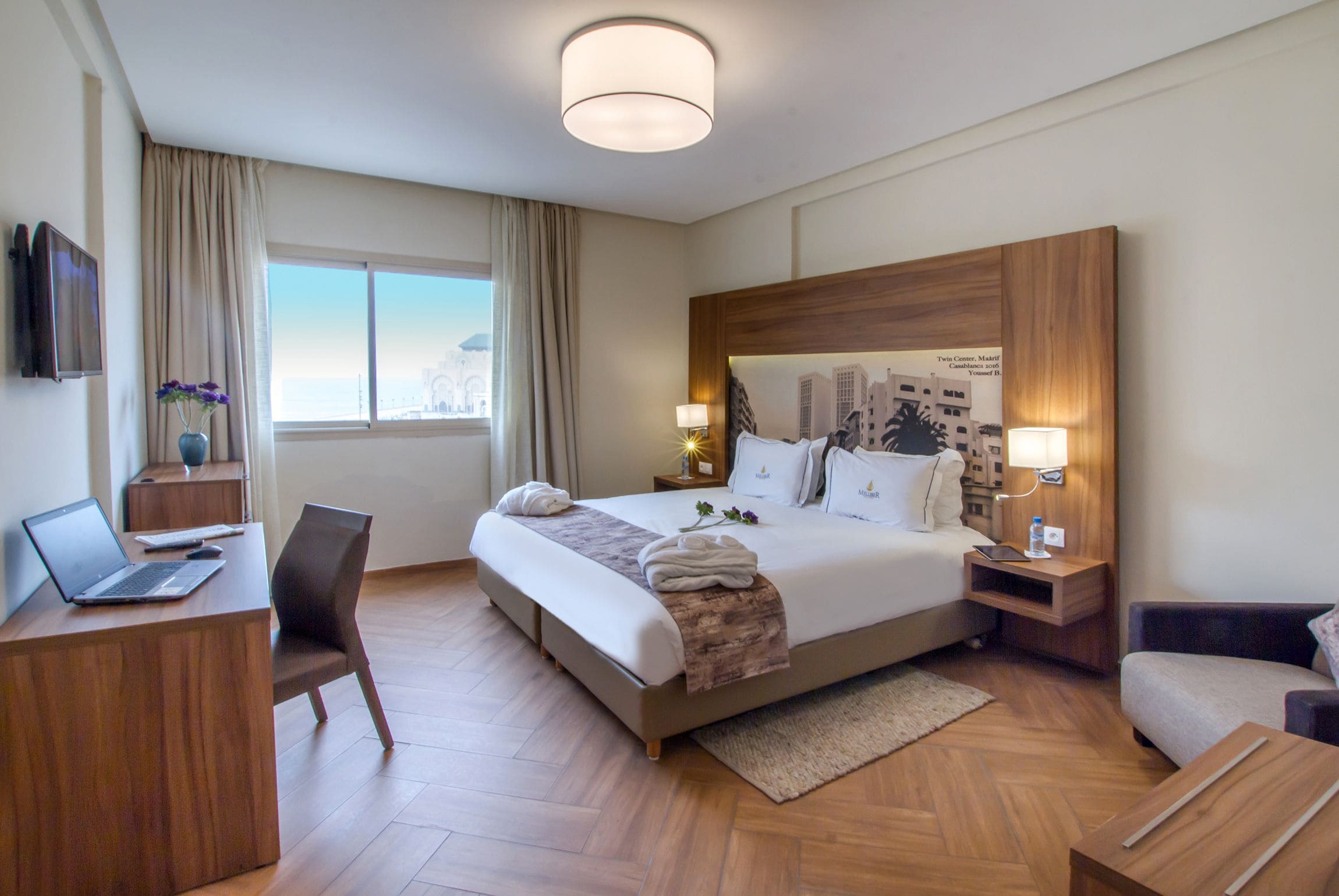 Habitación de hotel con una cama grande, un escritorio con una computadora portátil, una silla, un sofá y vista panorámica de la ciudad a través de la ventana.