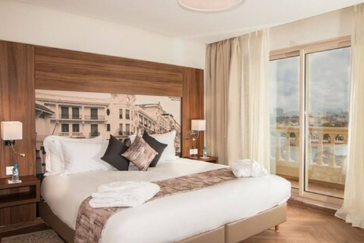 Ein Hotelzimmer mit einem großen Bett, einem Holzkopfteil, zwei Nachttischen mit Lampen, Blick auf die Stadt durch ein großes Fenster und einem Balkon mit Glasgeländer.