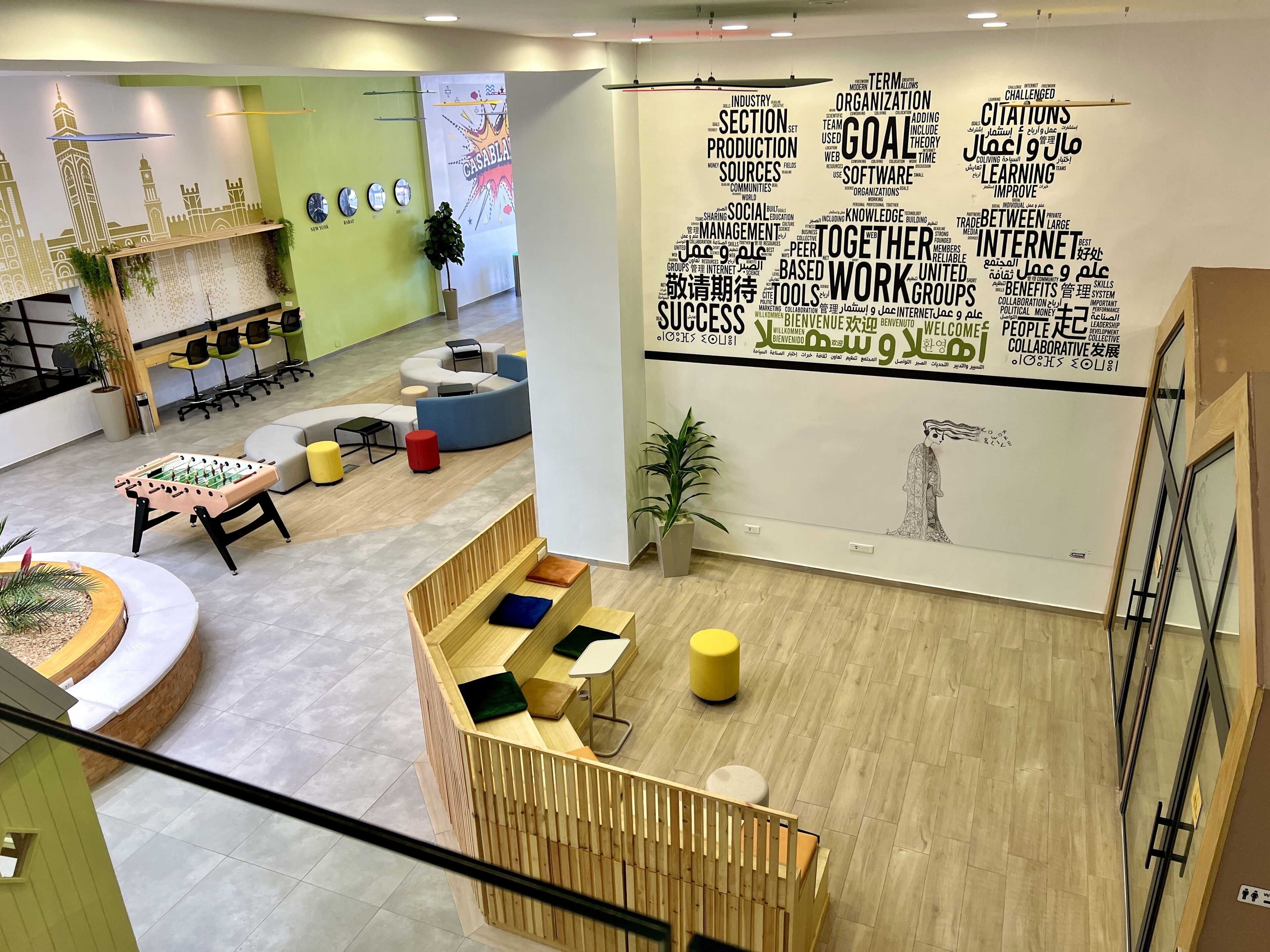 Ein moderner Arbeitsraum mit einer großen Wand mit motivierenden Wörtern und Sprüchen. Es gibt Sitzbereiche mit Sofas und Tischen, und ein Tischfußballtisch ist sichtbar.