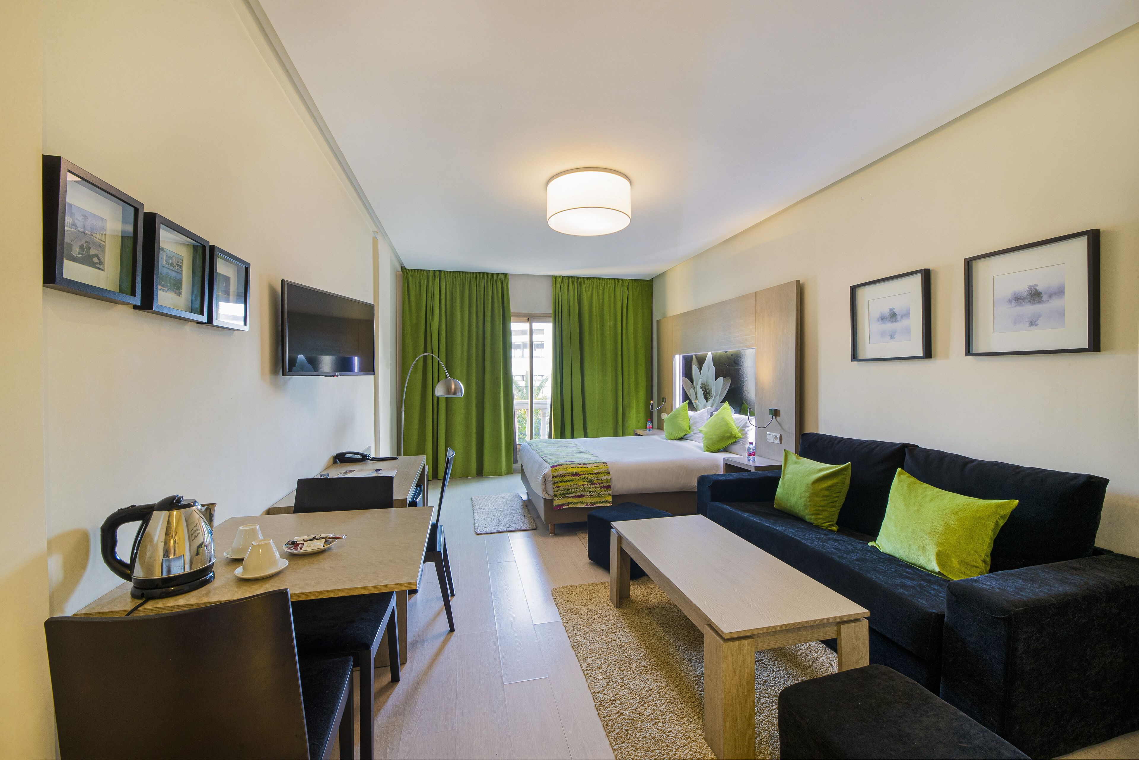 Habitación de hotel con cama, sofá, escritorio y mesa de centro. Se observan cuadros en las paredes y una ventana con cortinas verdes.
