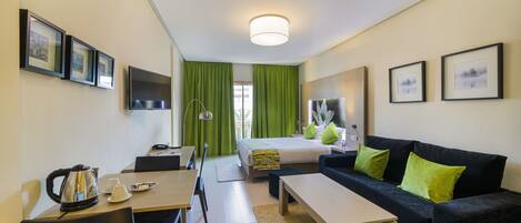 Habitación de hotel con cama, sofá, escritorio y mesa de centro. Se observan cuadros en las paredes y una ventana con cortinas verdes.