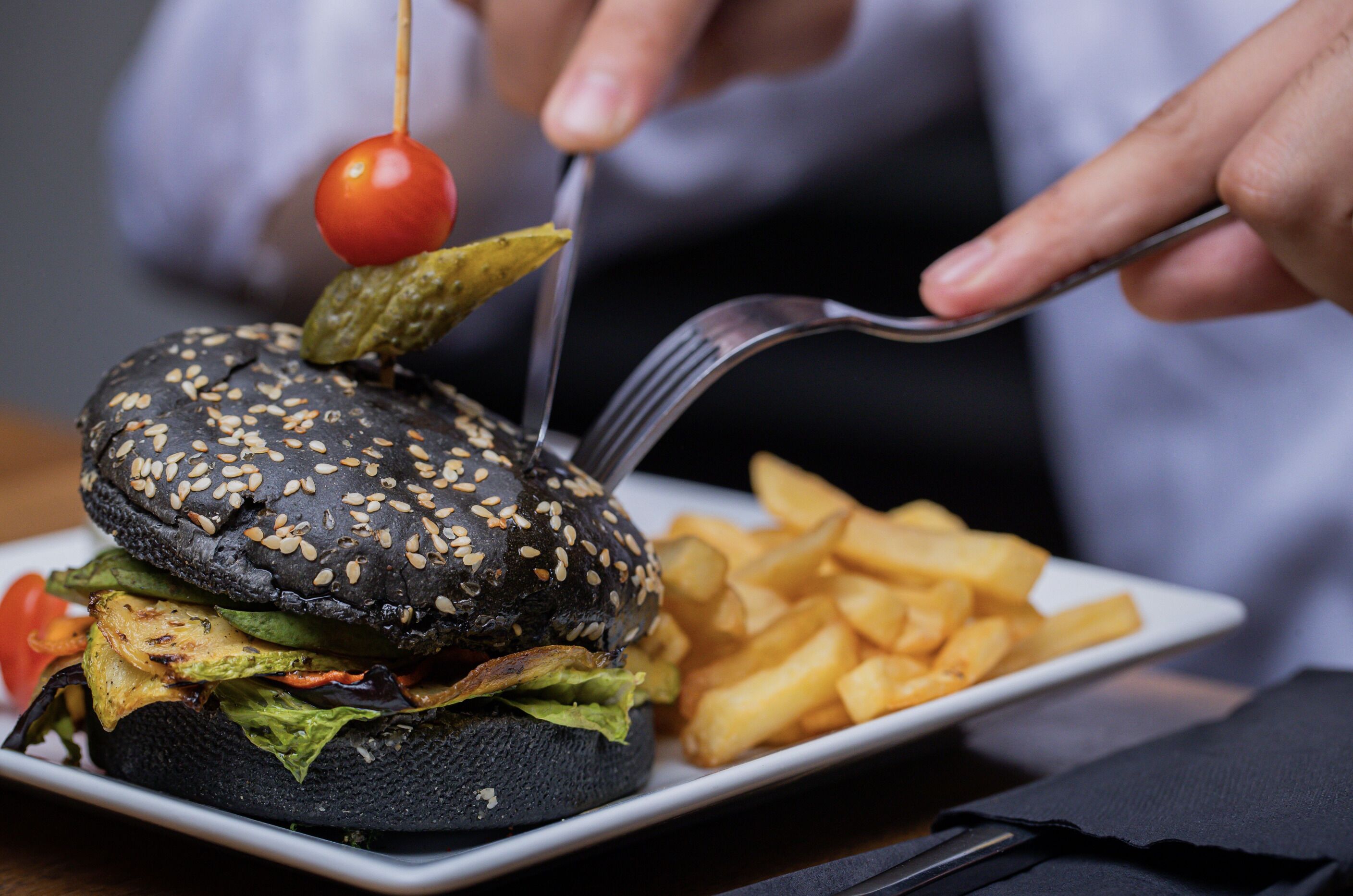 A black burger with sesame seeds, a cherry tomato, and pickles on a white plate, accompanied by French fries.
