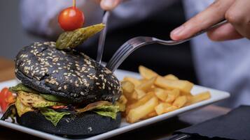 Una hamburguesa negra con semillas de sésamo, tomate cherry y pepinillos sobre un plato blanco, acompañada de papas fritas.