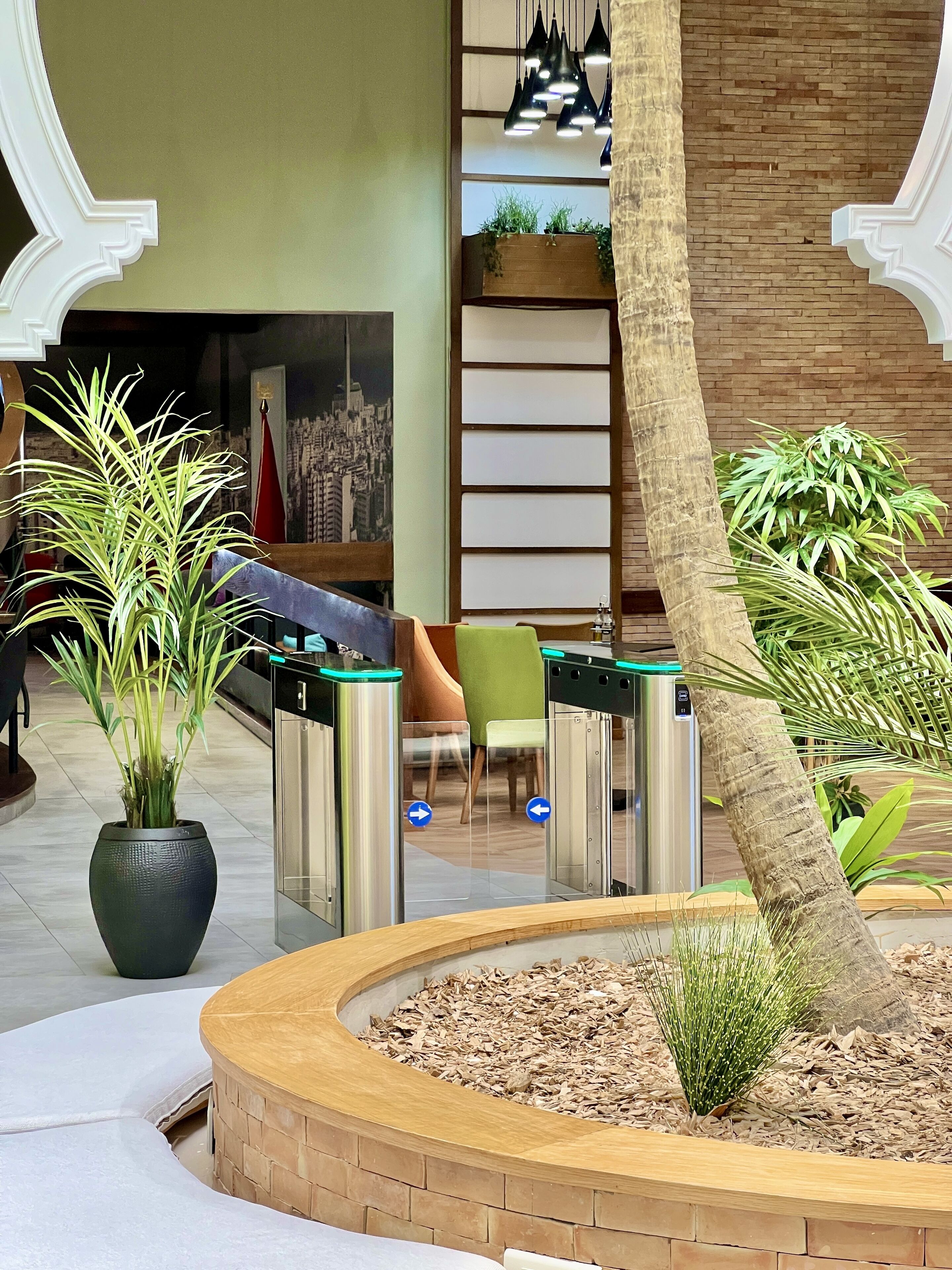 A lobby with a reception desk, a large potted plant, a wooden planter with a tree, and a brick wall with a cityscape mural.