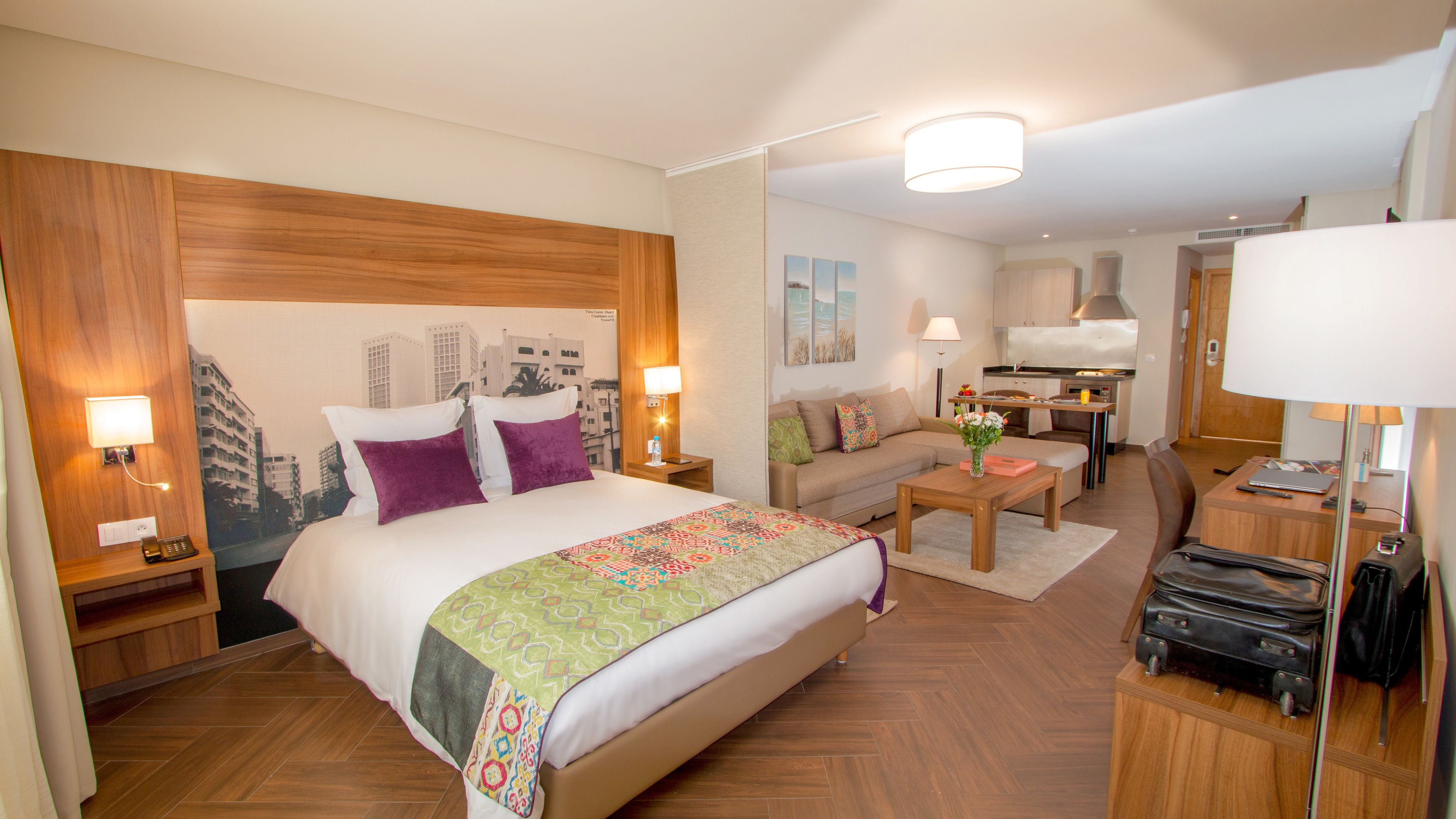 A hotel room with a large bed, a sofa, a small table, a desk, and a kitchenette.