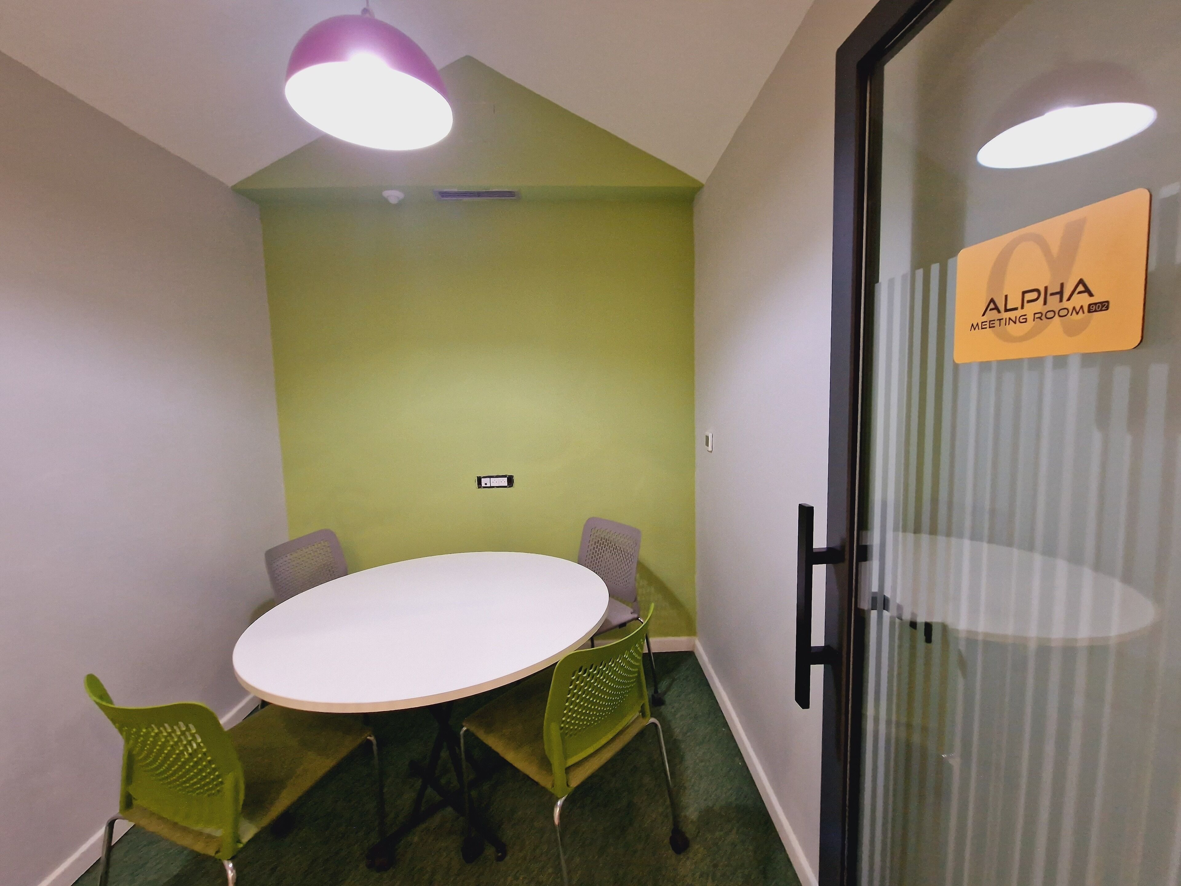 A meeting room with a white table, green chairs, and a glass partition with a sign reading "ALPHA MEETING ROOM 902".