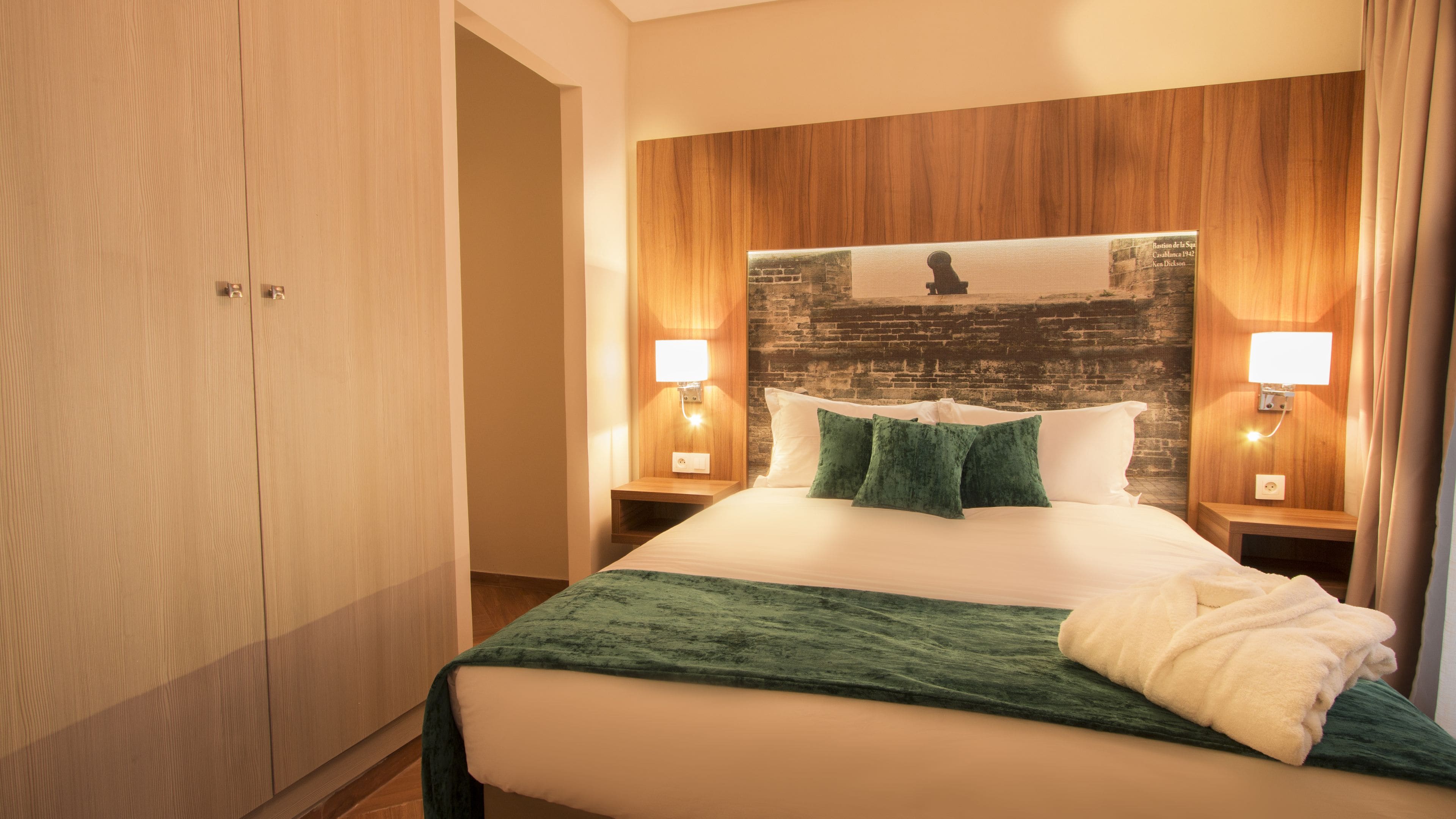 Habitación de hotel con una cama grande, cabecero de madera, dos mesitas de noche con lámparas y una manta verde.