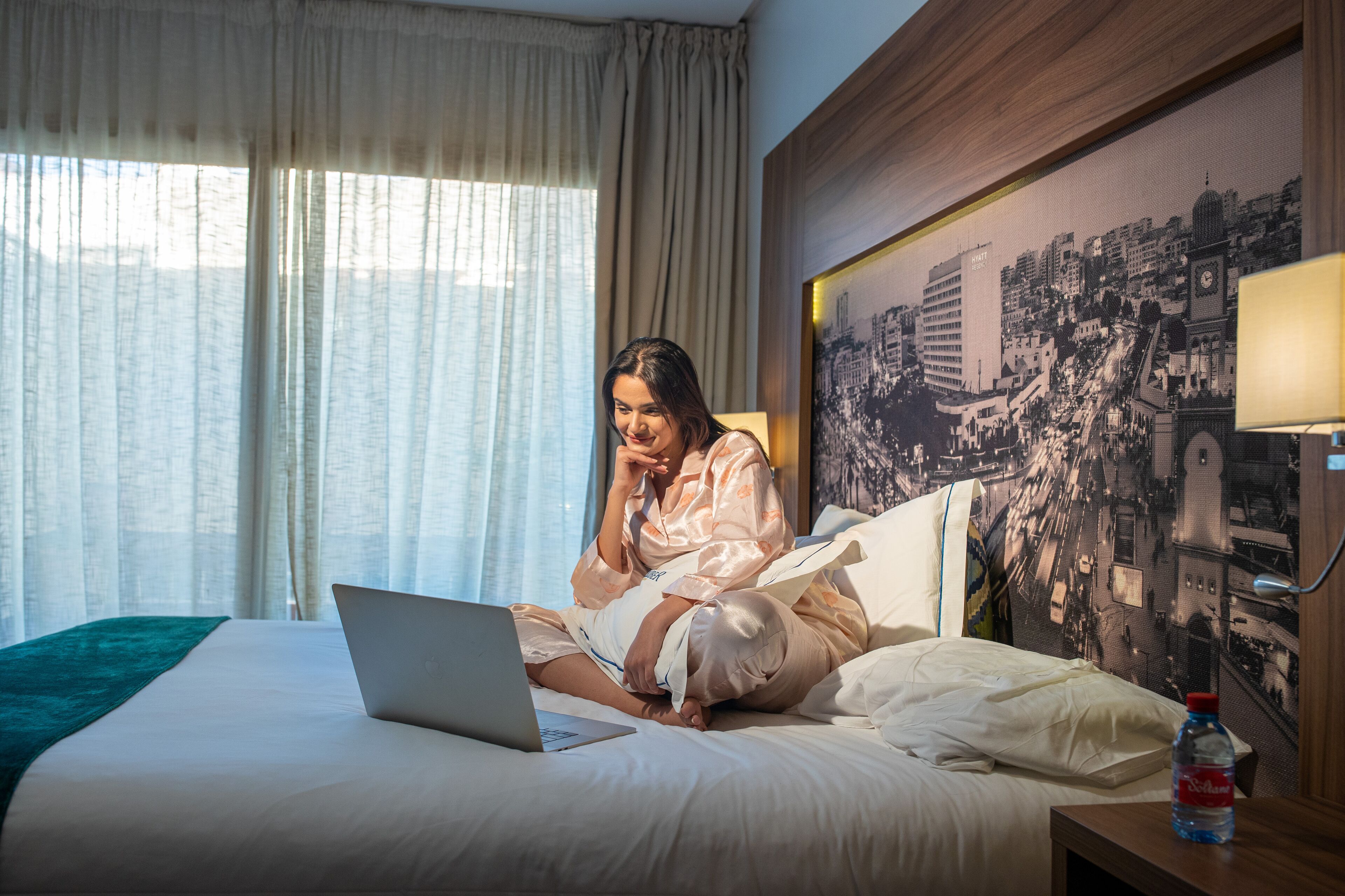 Une chambre d’hôtel avec un lit, un ordinateur portable, une bouteille d’eau et un tableau représentant un paysage urbain accroché au mur.